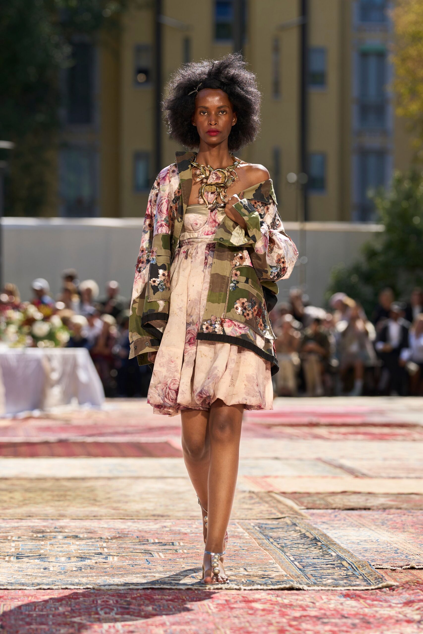 Oversized floral necklace, petals wired into a thorny bronze tangle. Dropped camouflage field jacket patched with sequined rose clusters. Blush floral bustier dress with a bubbled hem and one slipped strap. Barely there sandals, pearls marching up the toe strap. Walking across sun-warmed rugs toward lunch in a palace courtyard.