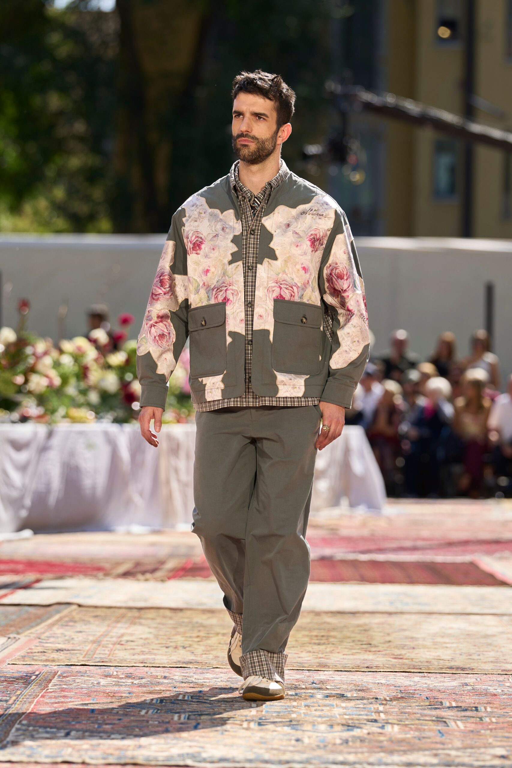 Sage utility jacket patched with watercolor rose panels and two flap pockets. Brown micro-check button-down peeking at the collar and dropped hem. Straight sage trousers with check lining turned up at the ankle. Cream trainers with tan rubber toe caps. Walking back from a garden lunch with pollen on your cuffs.