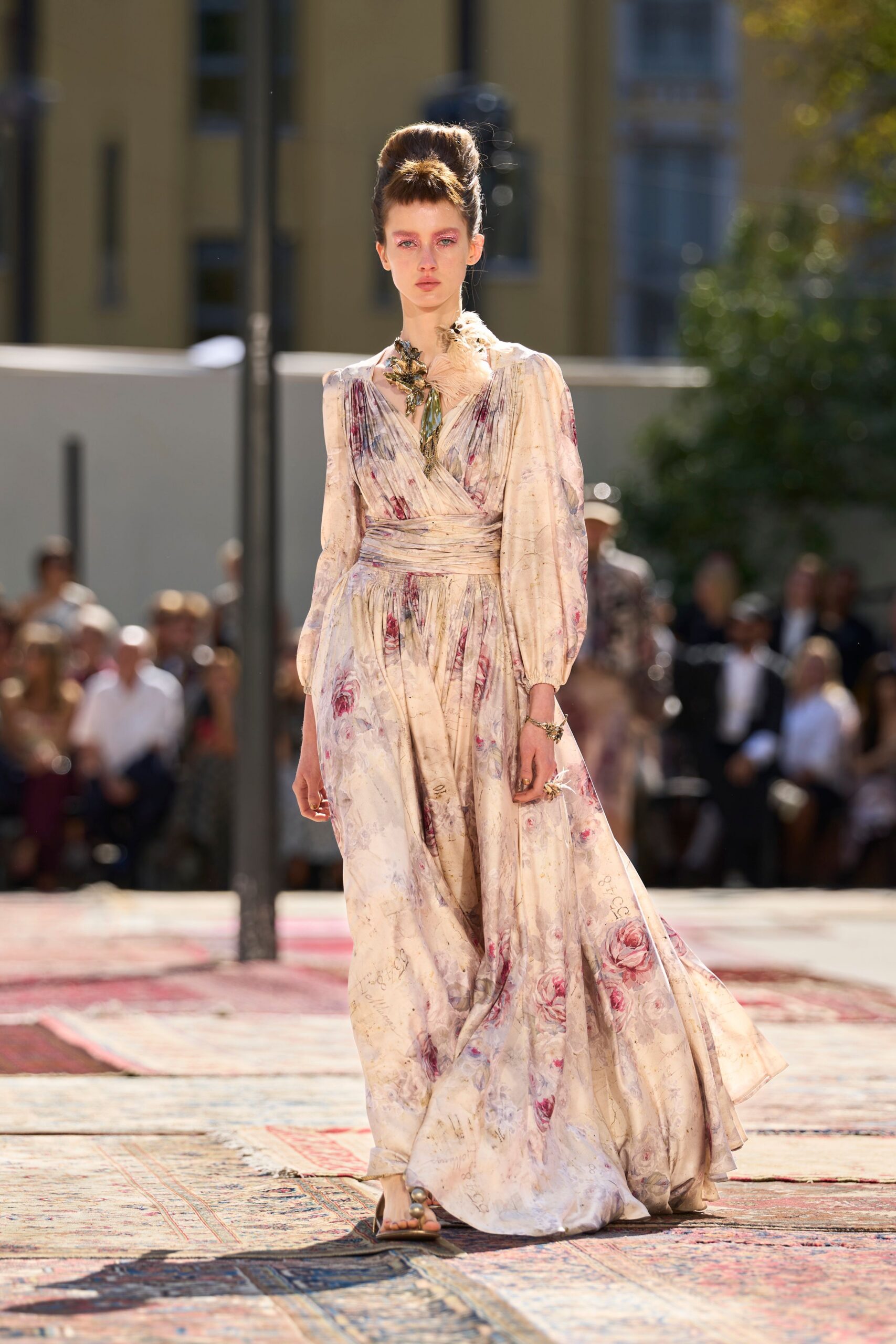 Blush silk maxi dress, rose print washed with faint handwritten script. Large feathered floral brooch pinned at the shoulder. Flat sandals with oversized pearl drops at the toes. Crossing a sunstruck courtyard after the garden ceremony has already started.