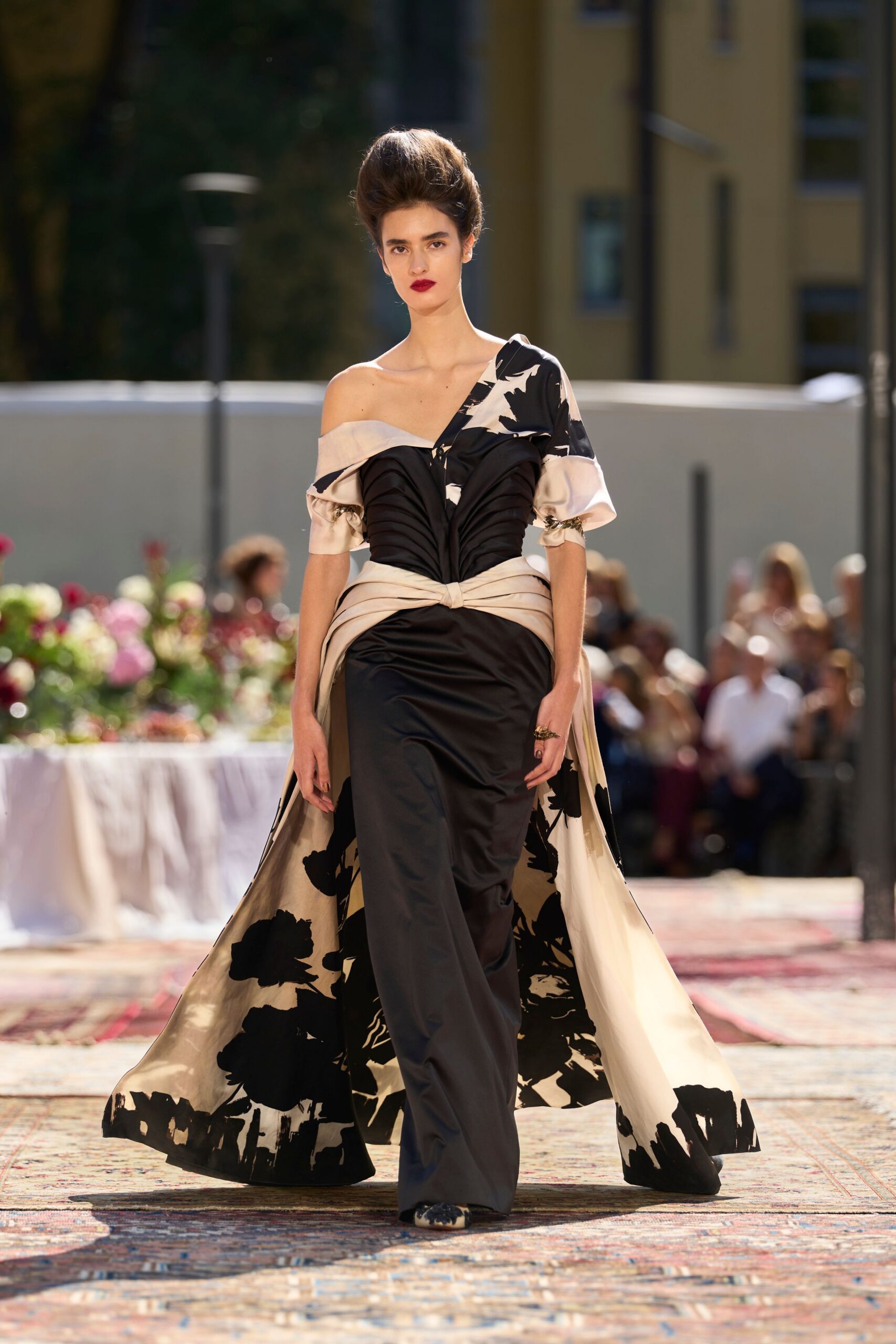 Black satin column gown with ruched bodice and knotted cream sash. Ivory overskirt printed with inky florals, falling open from the waist. Cream pumps with black applique at the pointed toe. Crossing a hotel courtyard after the wedding dinner has thinned out.