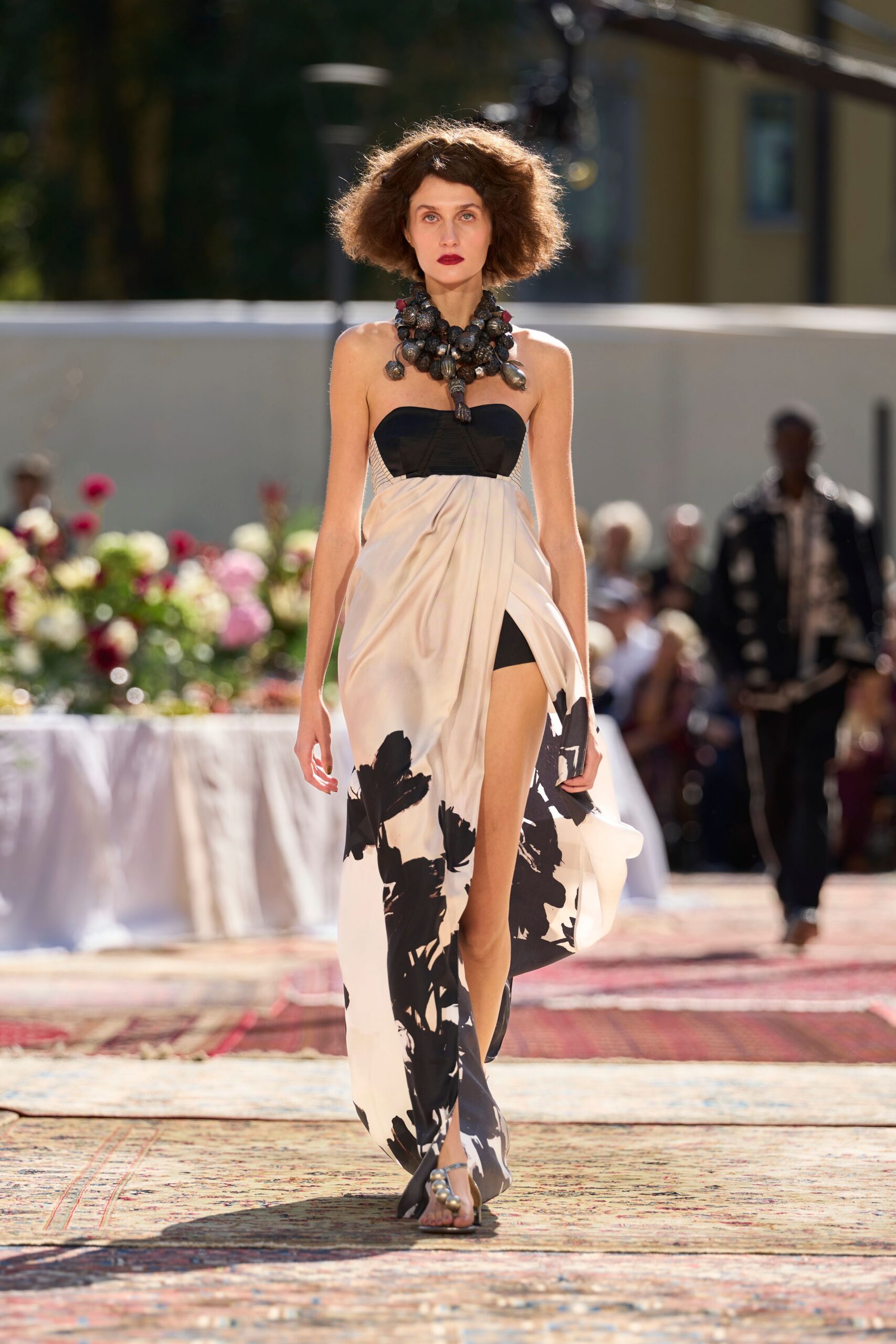 Clustered bead necklace with oversized charred-looking drops at the throat. Strapless ivory dress with a black bustier top and draped slit front. Black thong sandals with bulbous metallic beads across the toes. Garden luncheon after noon, white tablecloths still warm from the sun.