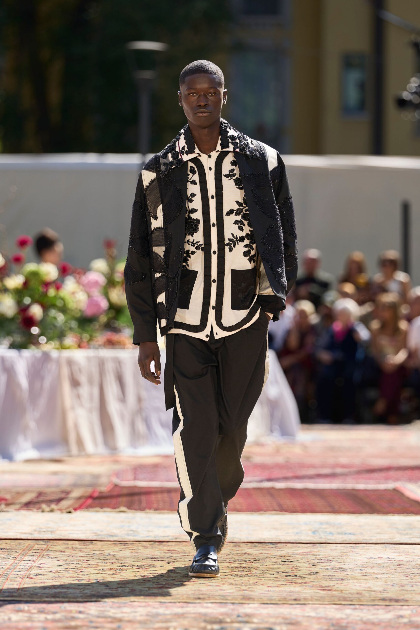 Black jacket overlaid with sheer panels and beaded leaf swirls. Ivory shirt traced with black ruffles and clustered floral beading. Black trousers with a chalk side stripe falling clean to the shoe. Square-toe black leather slip-ons. Garden luncheon running late, sun still hard on the paving.