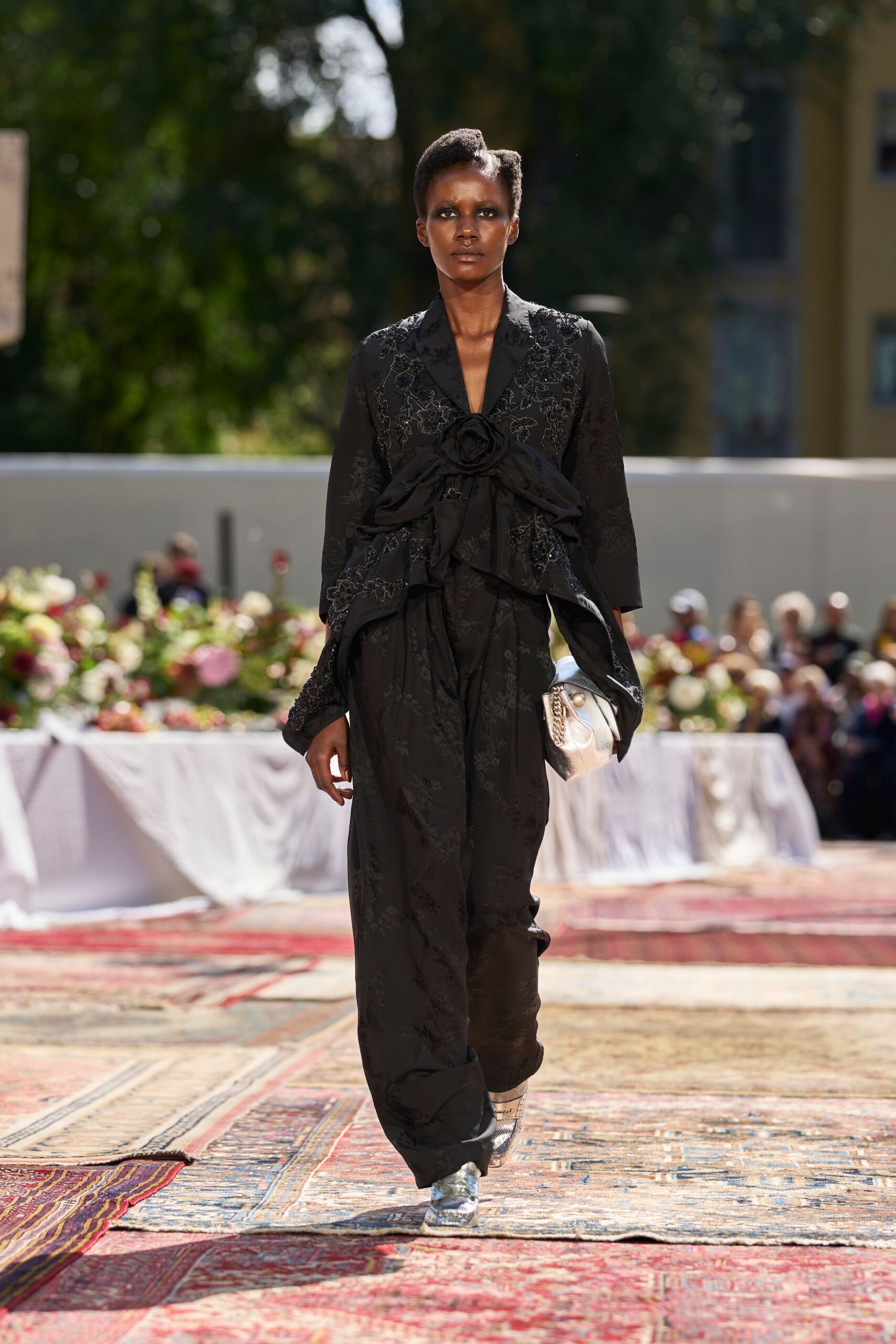 Black floral jacquard jacket, huge rosette bow cinched across the waist. Matching loose trousers pooling softly over the shoes. Silver clutch with a chunky chain draped at one side. Silver crinkled leather flats. Garden lunch under hard sun, white tablecloths lifting in the breeze.