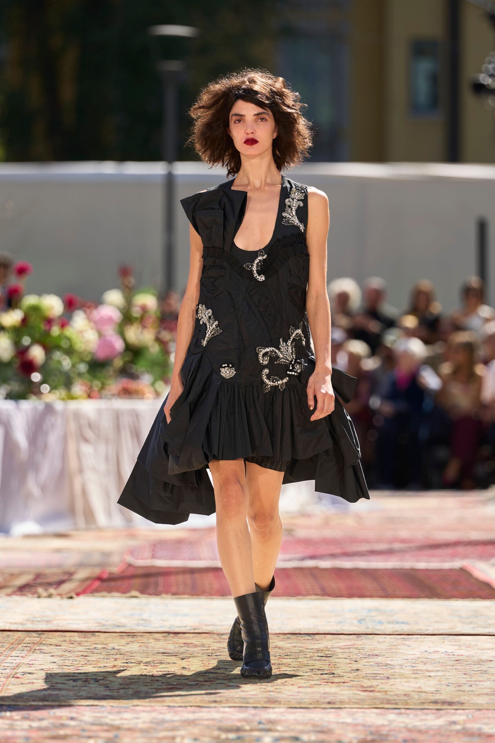 Black drop-waist dress in crisp taffeta, ruffled hem and silver beaded flourishes. Black ankle boots with softly slouched shafts. Garden luncheon after the speeches, sun catching the sequins.