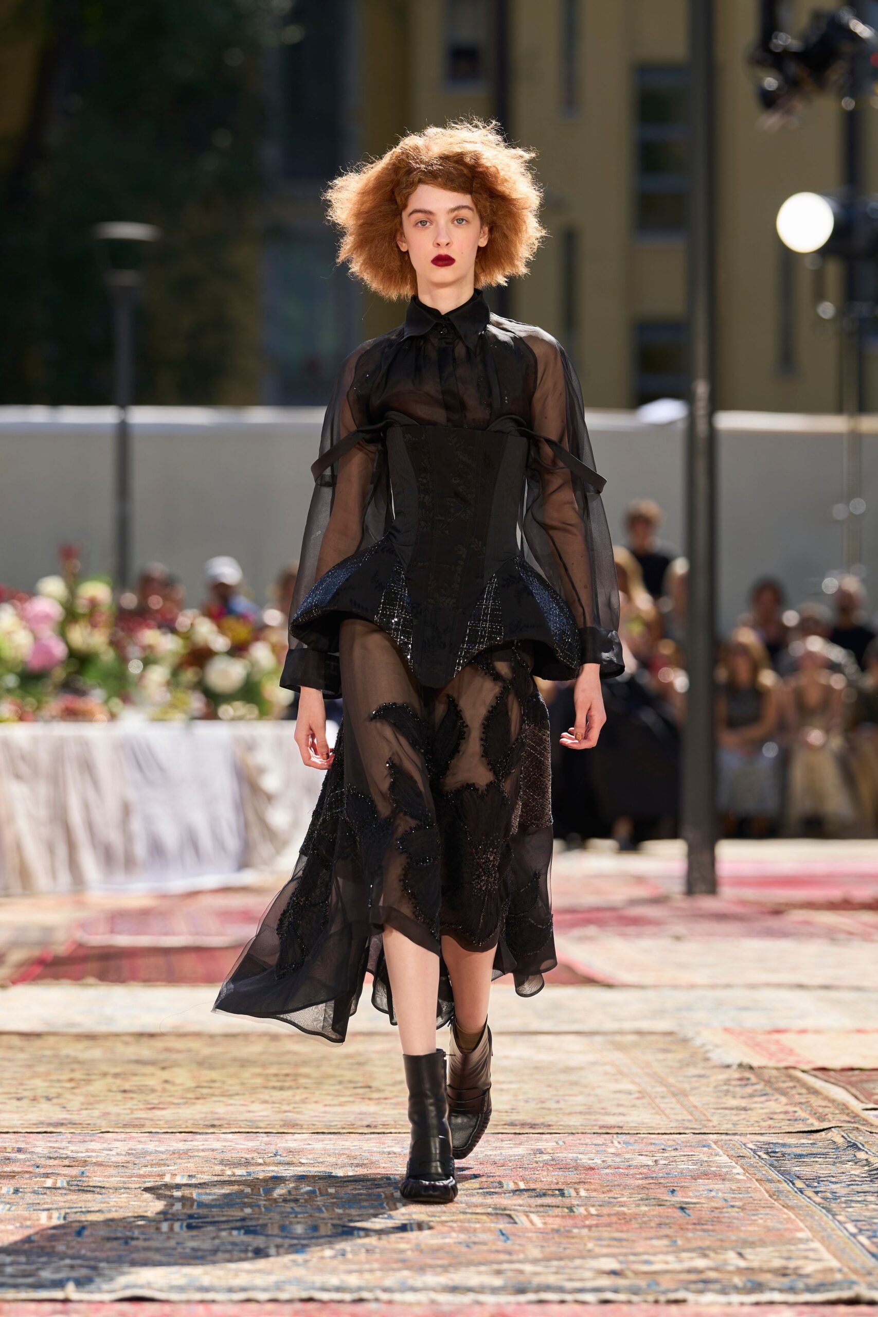 Black sheer organza blouse with a pointed collar and balloon sleeves. Long black corset panelled with faint floral brocade and glittered hip flares. Asymmetric sheer skirt scattered with black beaded leaf appliqué. Black ankle boots with square toes. Crossing a hotel courtyard after the wedding lunch has gone cold.