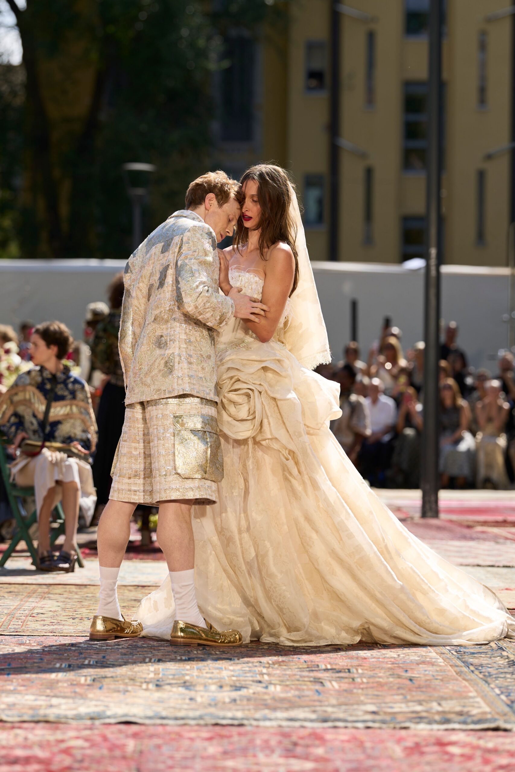 Soft ivory tulle veil falling past the shoulder in a sheer panel. Strapless ivory gown with a ruched bodice and enormous rosette hip drape. Sun-struck vows on a courtyard carpet, guests fanning themselves through the heat.