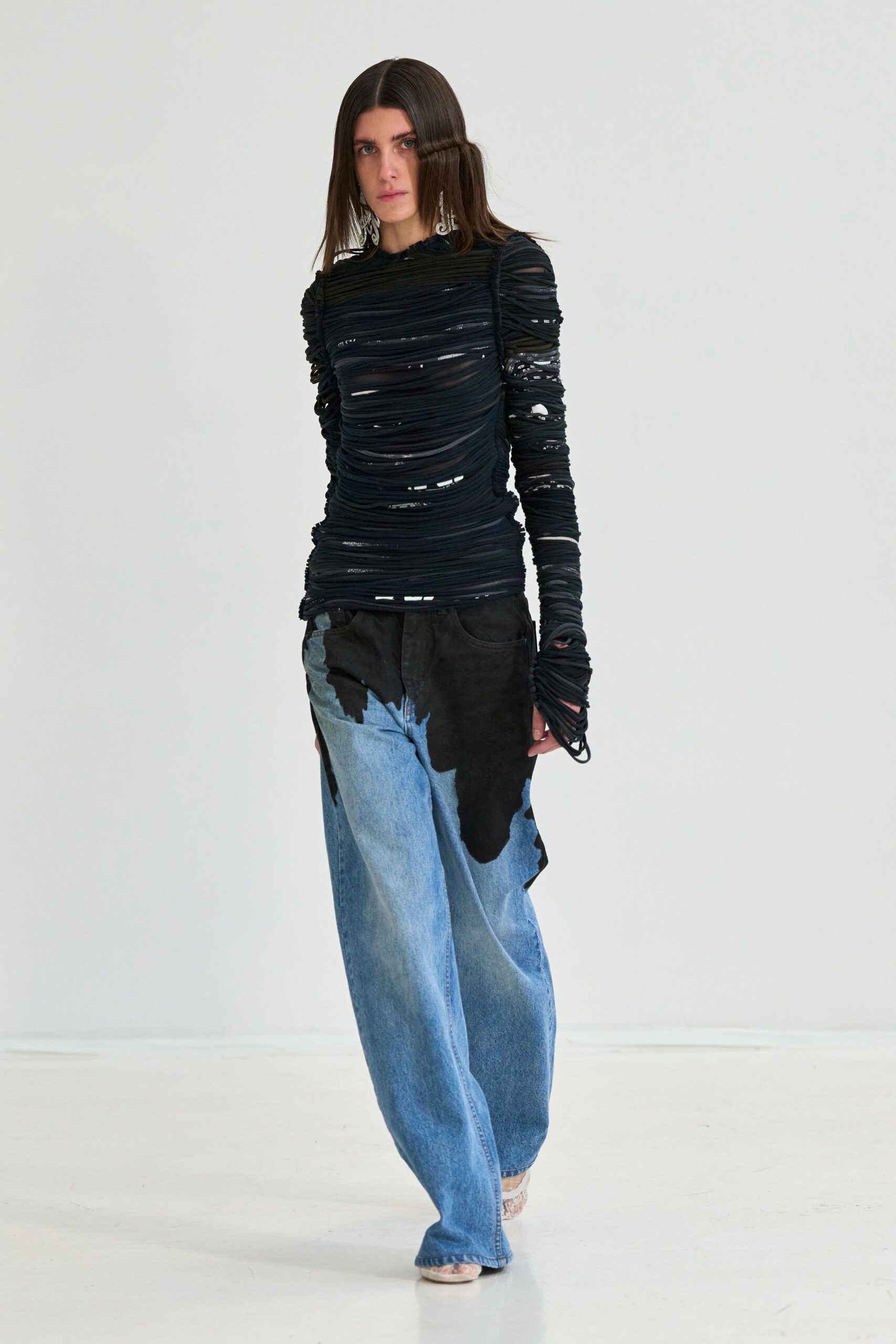 Long silver filigree drops brushing the jaw. Black ruched mesh top, slashed into narrow bands across the body. Pale wide-leg denim with irregular black overdye pooling from the waistband. Snakeskin peep-toes just visible under the hem. Walking from a warehouse party into the first washed-out morning light.