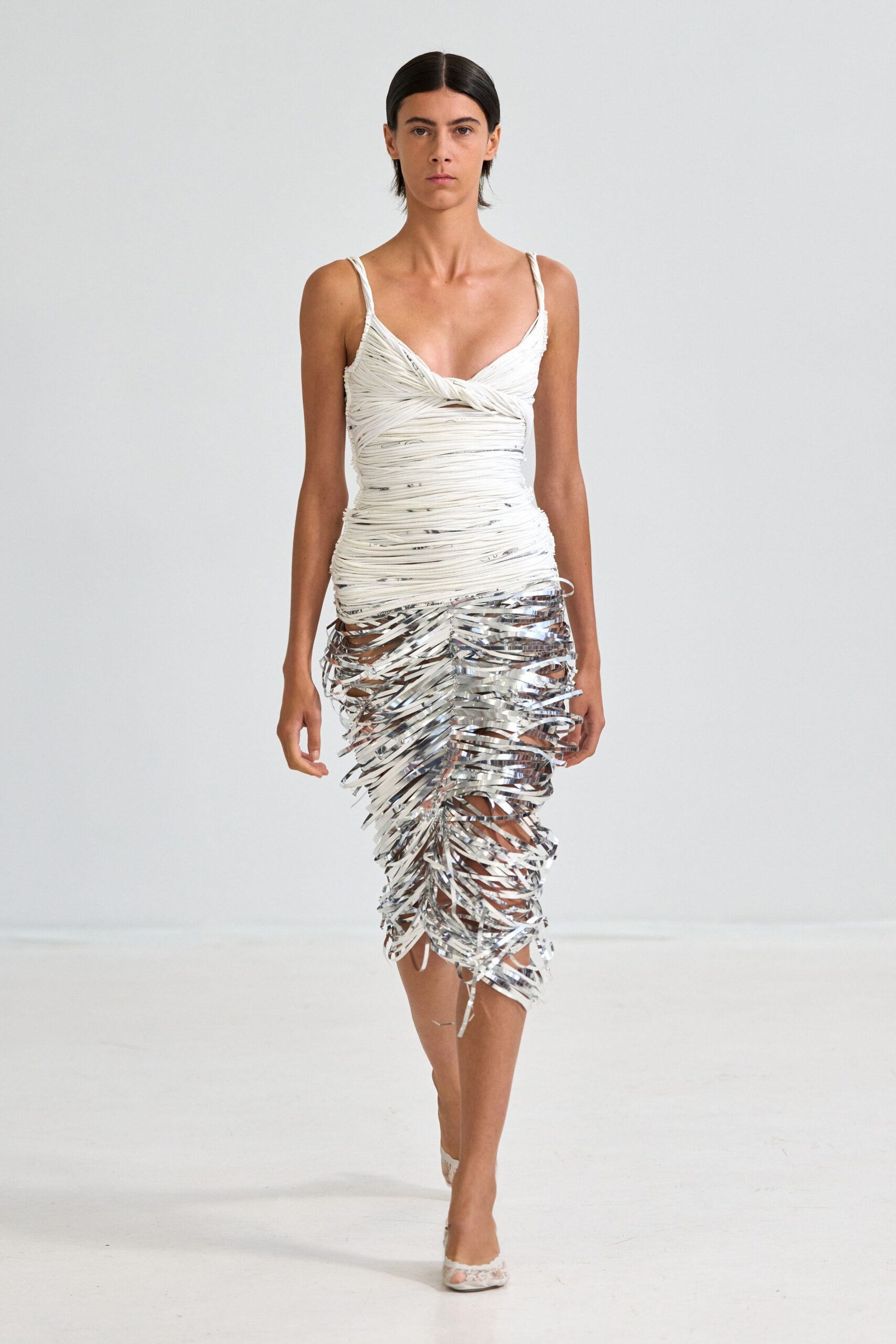 Ivory spaghetti-strap dress, bodice wrapped in narrow cords knotted at the bust. Silver foil streamers woven open, trailing in uneven strips below the hip. White lace peep-toe heels. Whatever catches the flash best at a rooftop birthday after midnight.