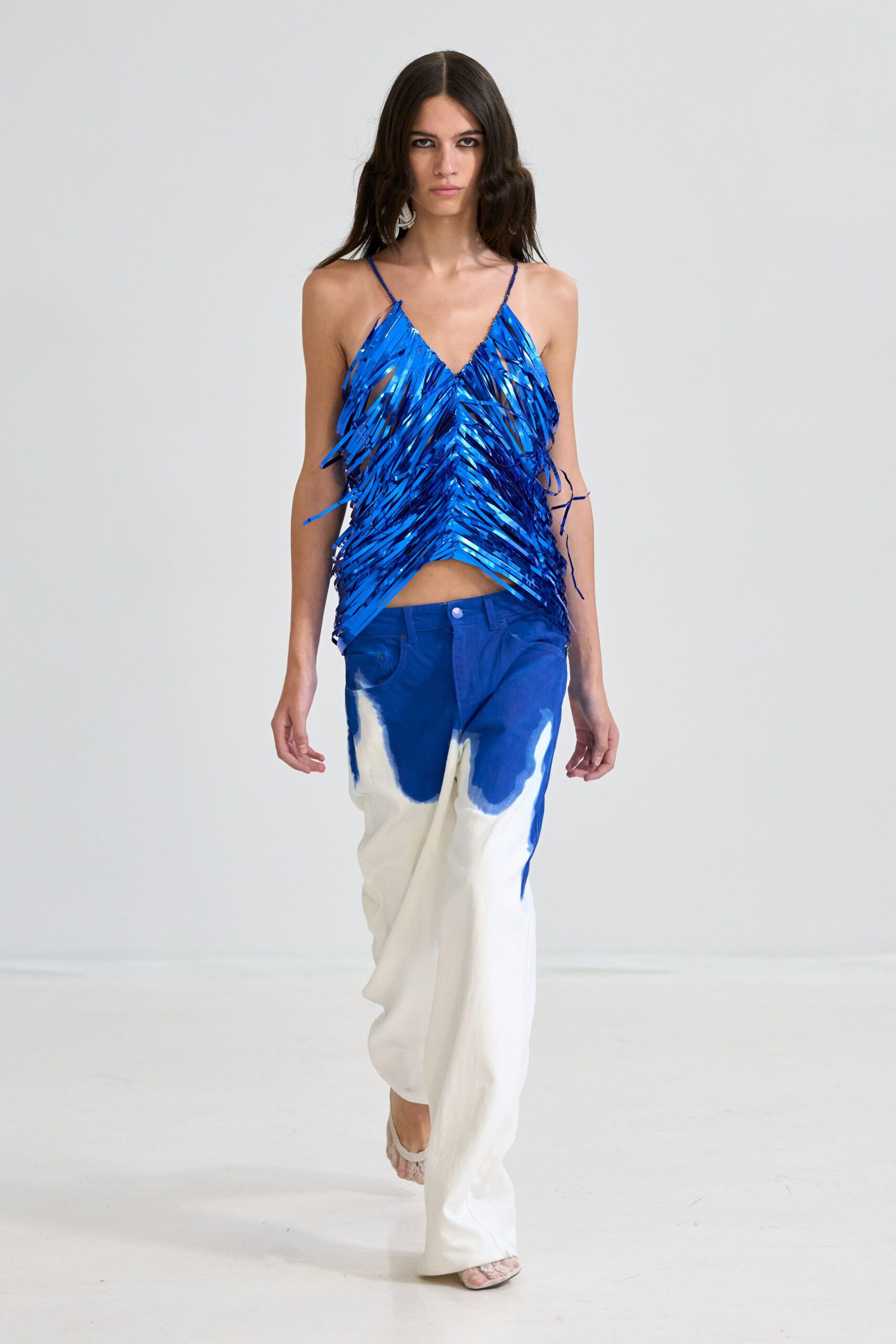 Cobalt strip-fringe camisole, glossy ribbons crossing into a pointed hem. White wide-leg trousers dipped in cobalt, rivet jeans waistband peeking through. Pale snakeskin mules. Walking the harbor promenade after rain, blue paint still wet on the railings.