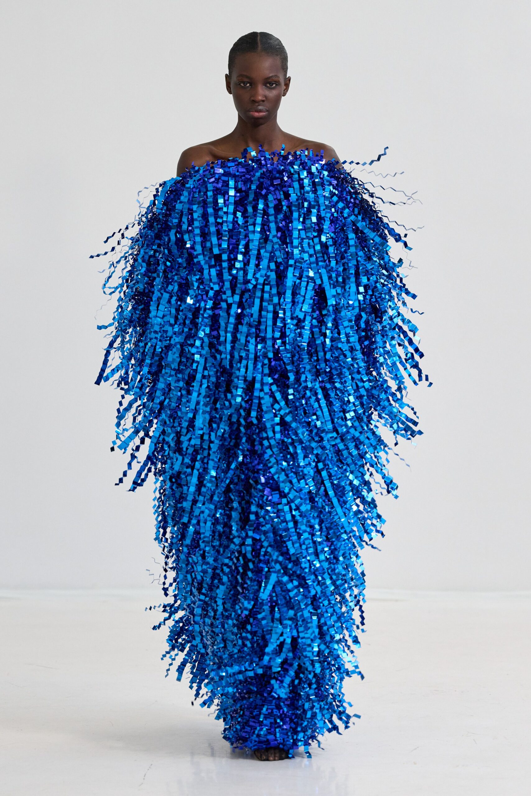 Off-the-shoulder column gown buried under electric-blue tinsel strips, stray curls kicking outward at the sleeve line. Crossing a parade route after midnight while the confetti is still stuck to everything.