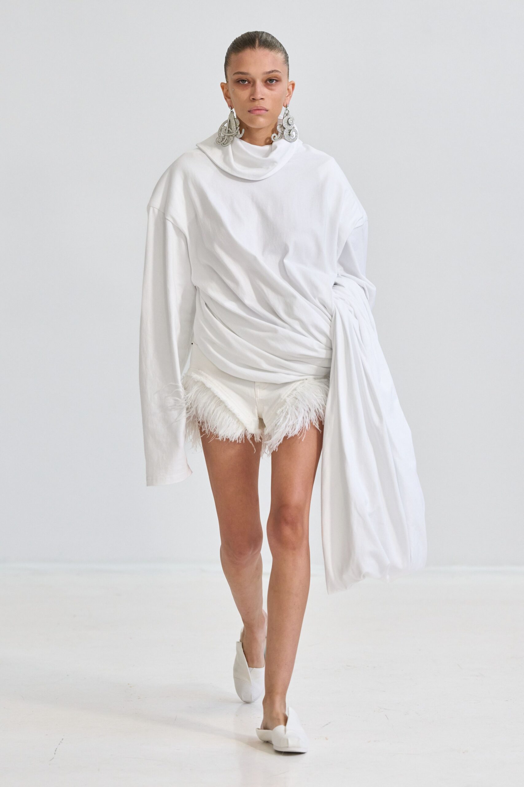 Oversized silver filigree drops curling almost to the collarbone. White jersey top with a rolled cowl and one sleeve hanging floorward. White shorts trimmed in shaggy feather fringe peeking under the hem. White folded-leather mules with blunt squared toes. Hotel hallway at dawn, carrying yesterday's sheets to the laundry room.