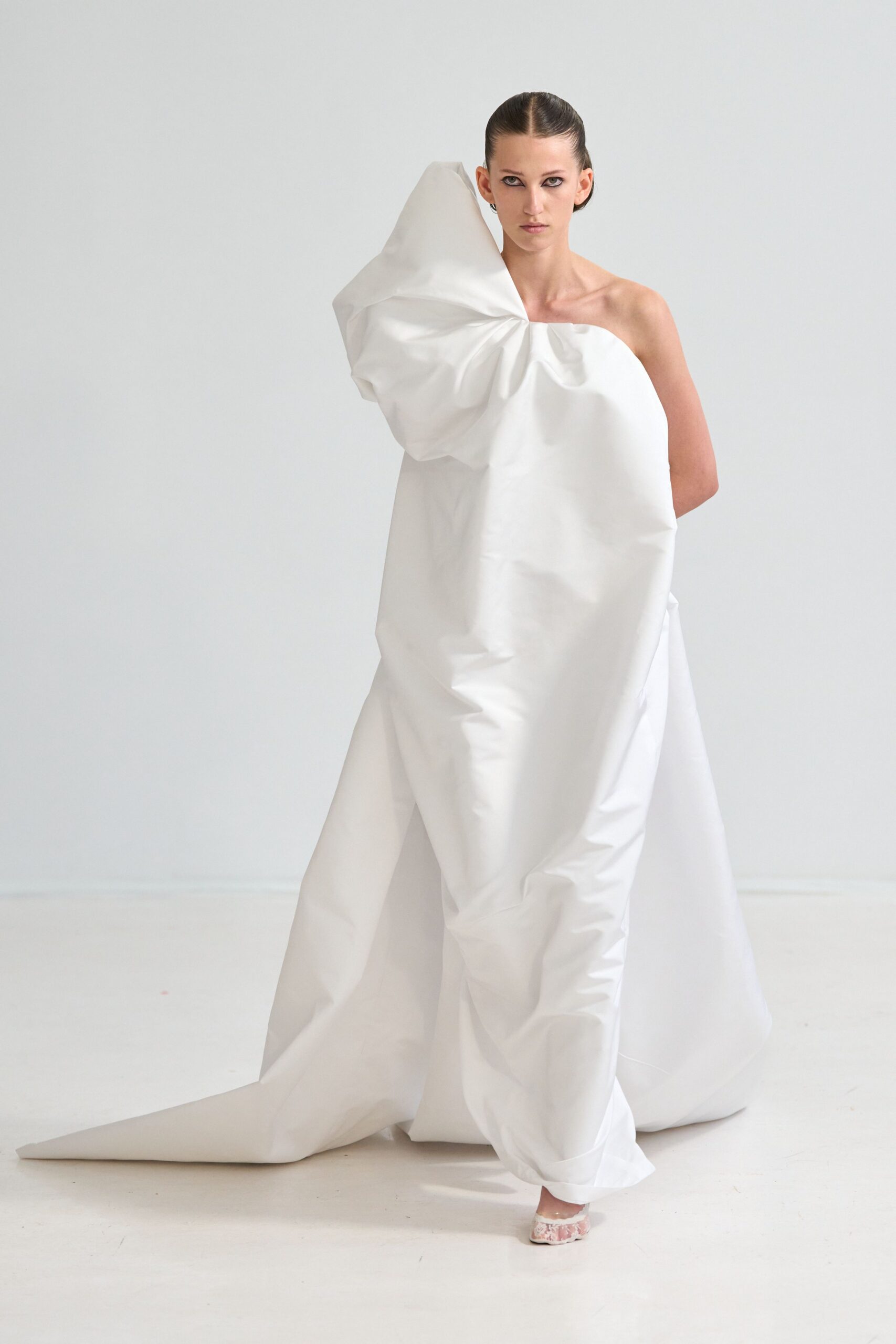 White sculptural one-shoulder gown in crisp paperlike cloth, knotted into a giant shoulder swell. Pale pink floral brocade peeping from the pooled hem. Crossing a silent gallery while the plaster dust still hangs in the air.