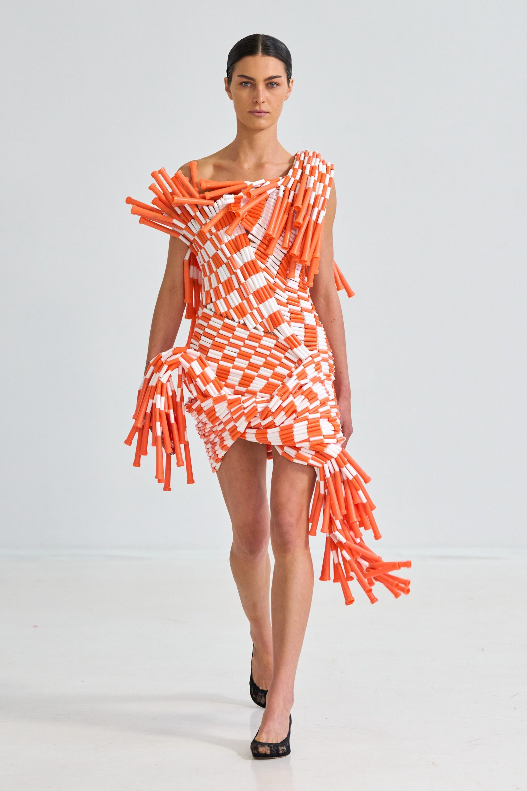 Asymmetric mini built from orange-and-white traffic batons, one shoulder spiking outward. Black lace pumps, sheer at the toe. Crossing a blocked-off street before the parade barriers come down.