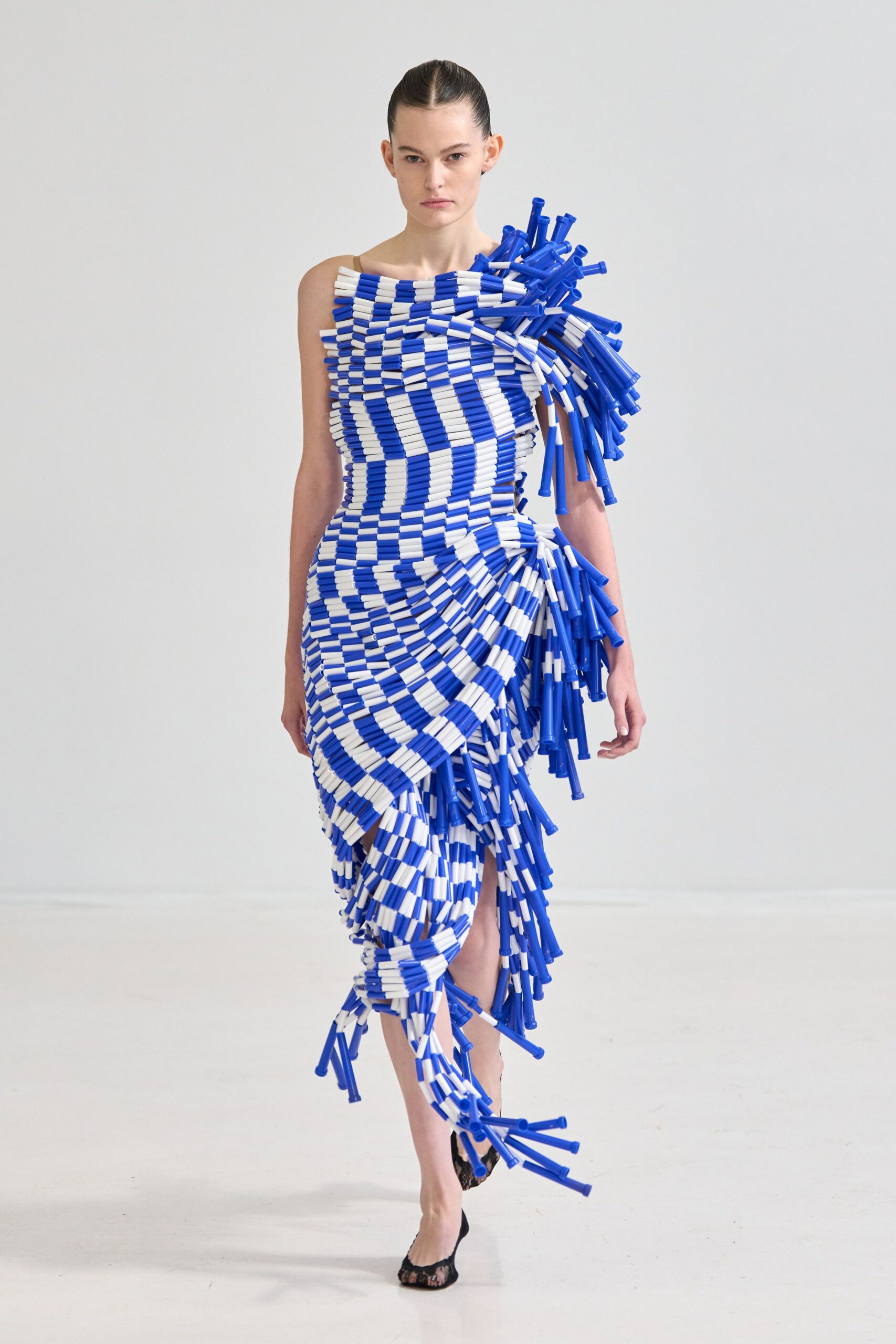 One-shouldered dress built from blue and white plastic tubes, spiraled into dense ridges. Black lace pumps, peep toe opening just visible. Crossing a ferry deck with salt drying on the rail.