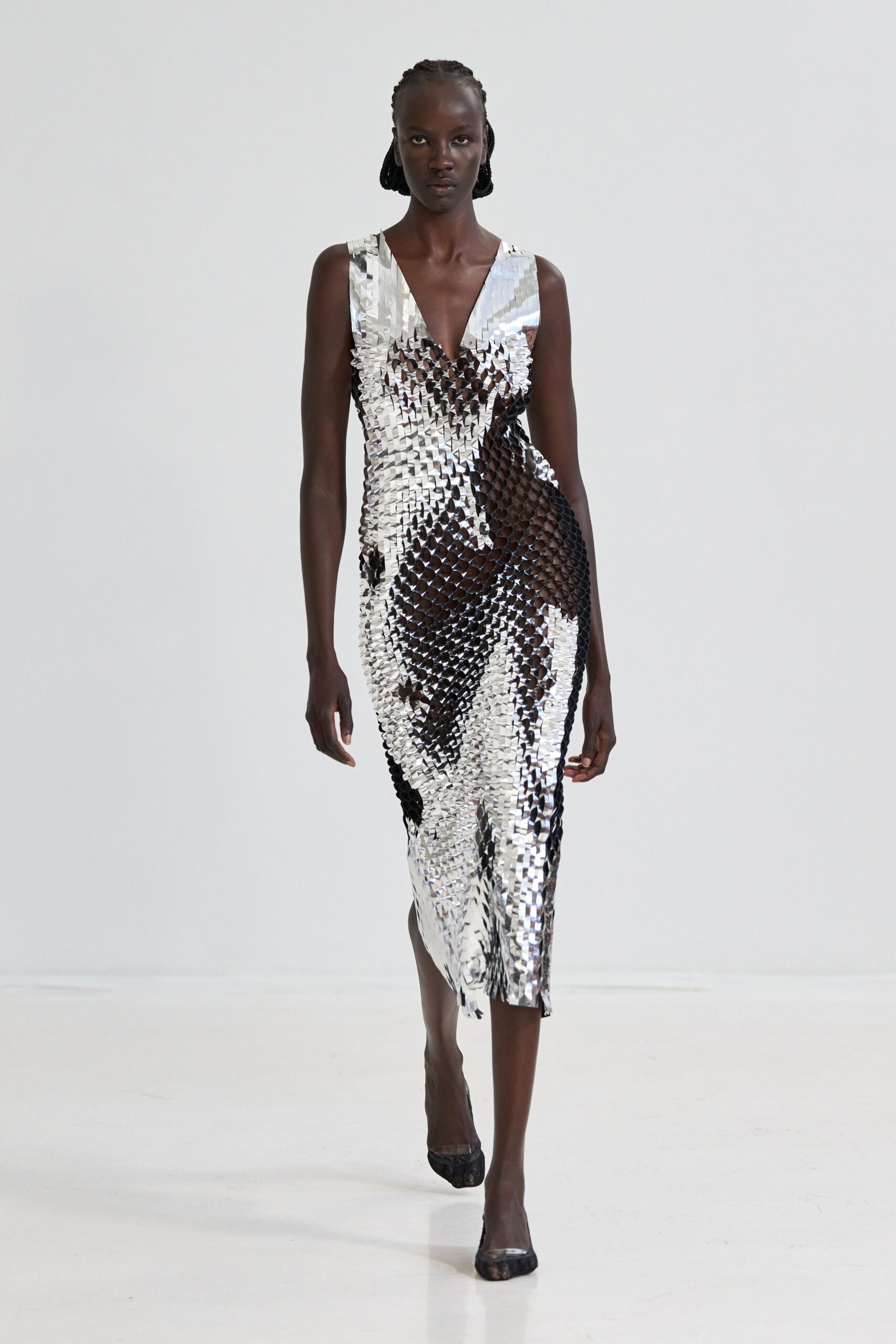 Mirror-paillette sheath dress, plunging V and a raw fringe at the slit. Black mesh pumps with a clear vinyl cap. Crossing a hotel atrium while the florist is still hosing the stone floor.