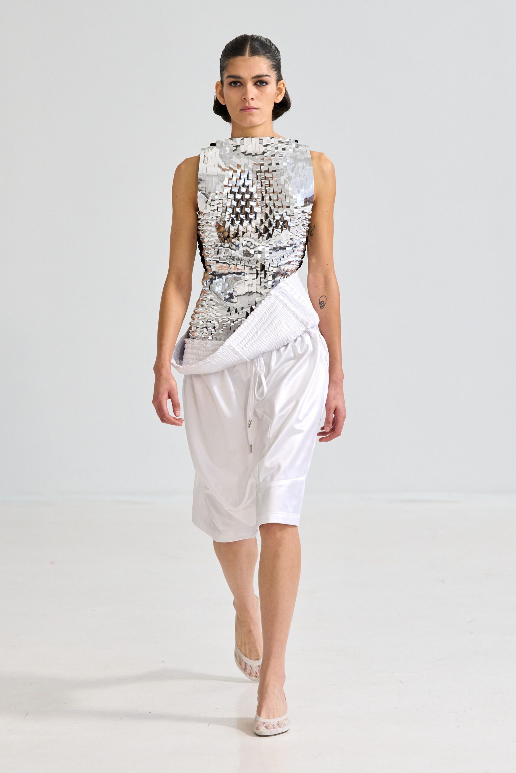 Sleeveless silver top built from tiny mirrored tabs, sides left open. White knee-length shorts in glossy nylon, drawcord tips hanging long. Quilted white tube wrapped low, curling up from one hip. Pale pumps with a cloudy mesh toe. Walking past a hotel ice machine in borrowed clothes after sunrise.
