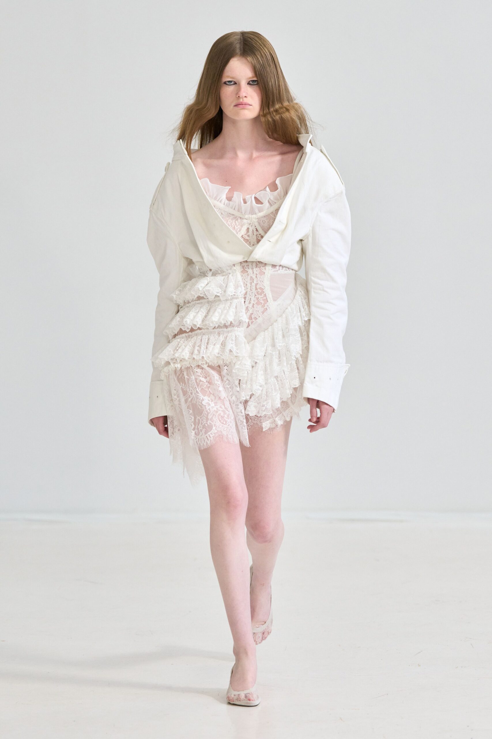 Off-white cotton jacket hanging open, collar points kicked sharply upward. Sheer ivory lace slip with tiered ruffles knotted at one hip. Pale lace-covered pumps with a low curved topline. Morning after the wedding, still barefoot in your head.