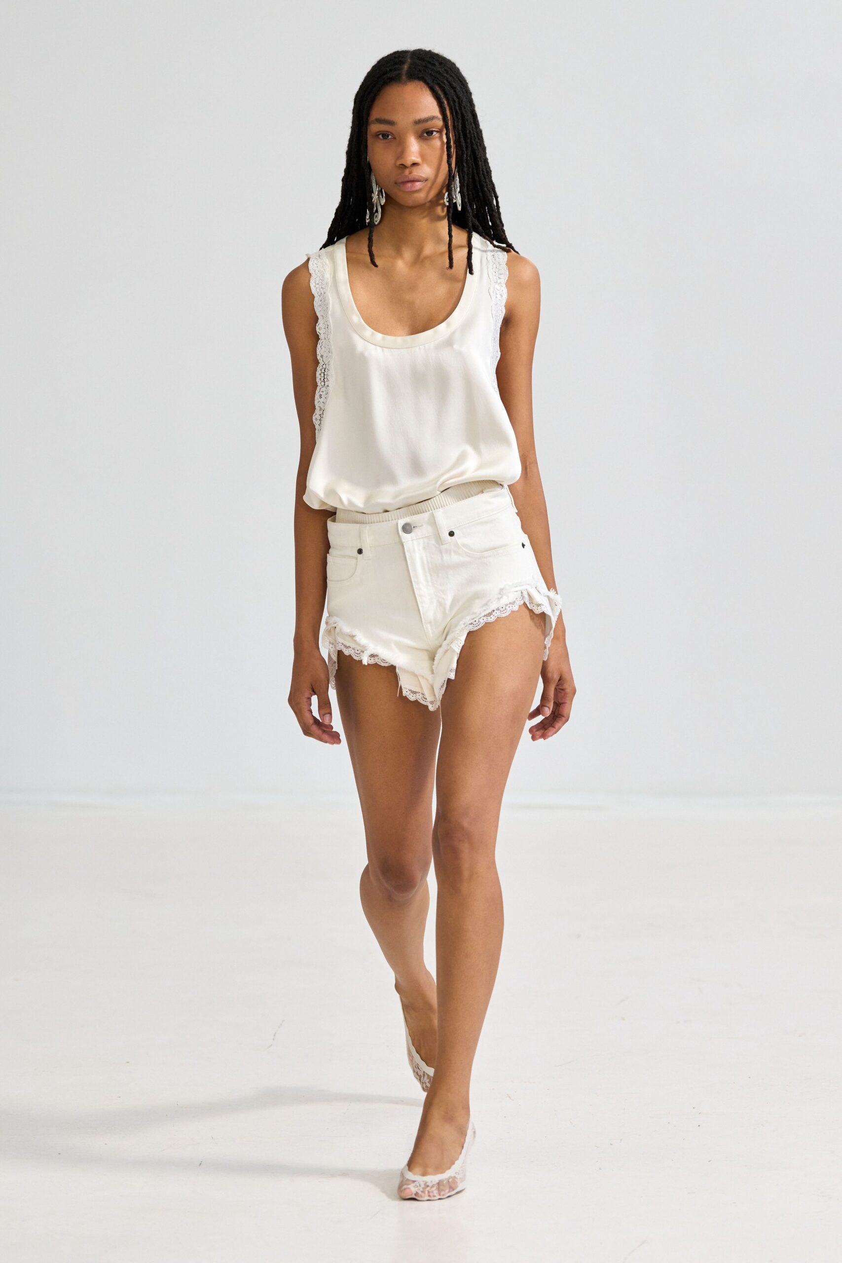 Ivory satin tank with scalloped lace running down the side seams. Low-rise white denim shorts, lace edging the curled cutaway hems. White lace mules with a clear peep-toe vamp. Long silver drops, leaflike and faceted. Morning after a beach wedding, crossing the rental house barefoot except for heels.