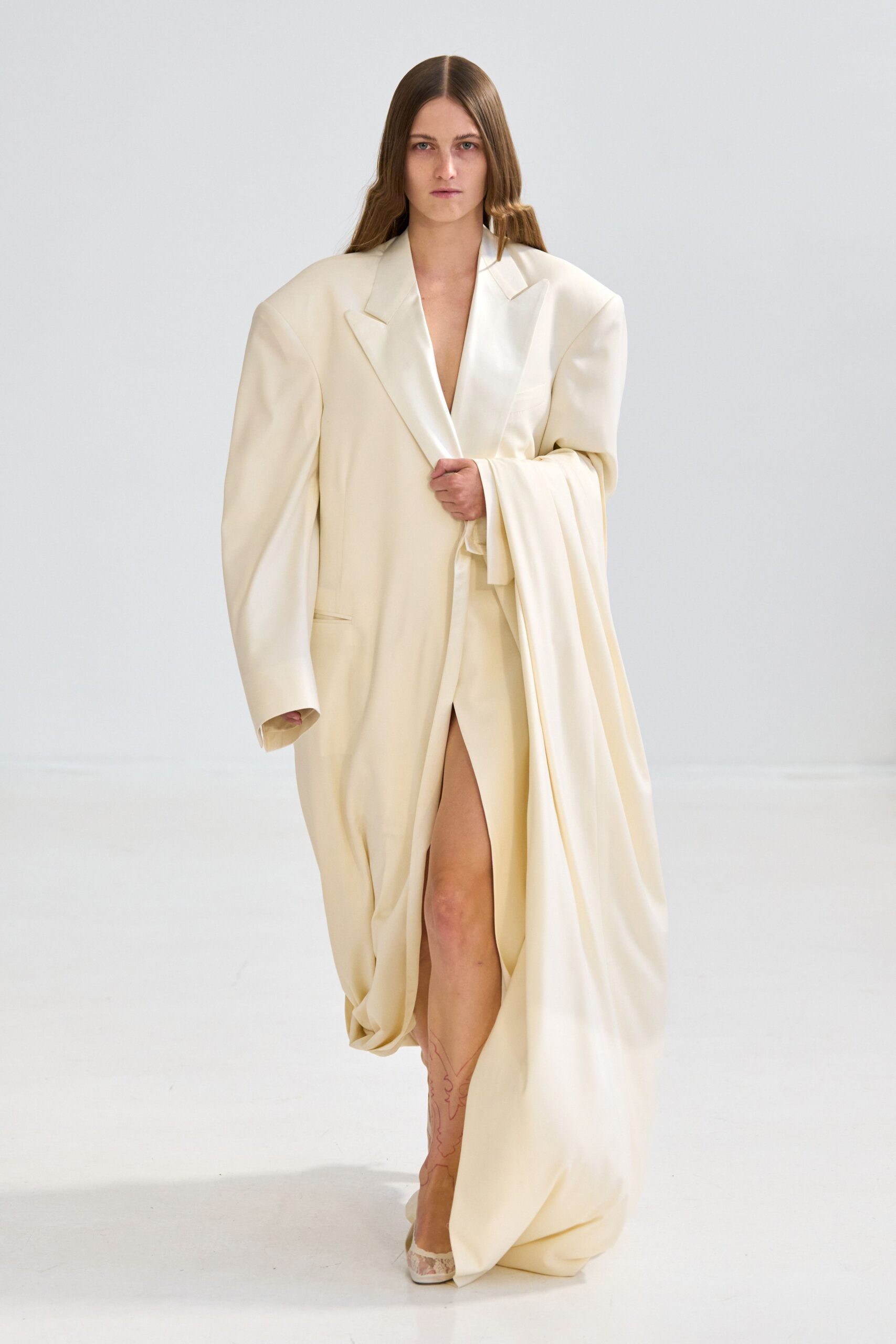 Ivory tailored coatdress with hard padded shoulders and a deep bare plunge. Heavy cream panels swept over one arm, pooling into a twisted hem. Pointed lace heels peeking out beneath the dragging front slit. Whatever you wear home after the civil ceremony turned into an all-night hotel stay.