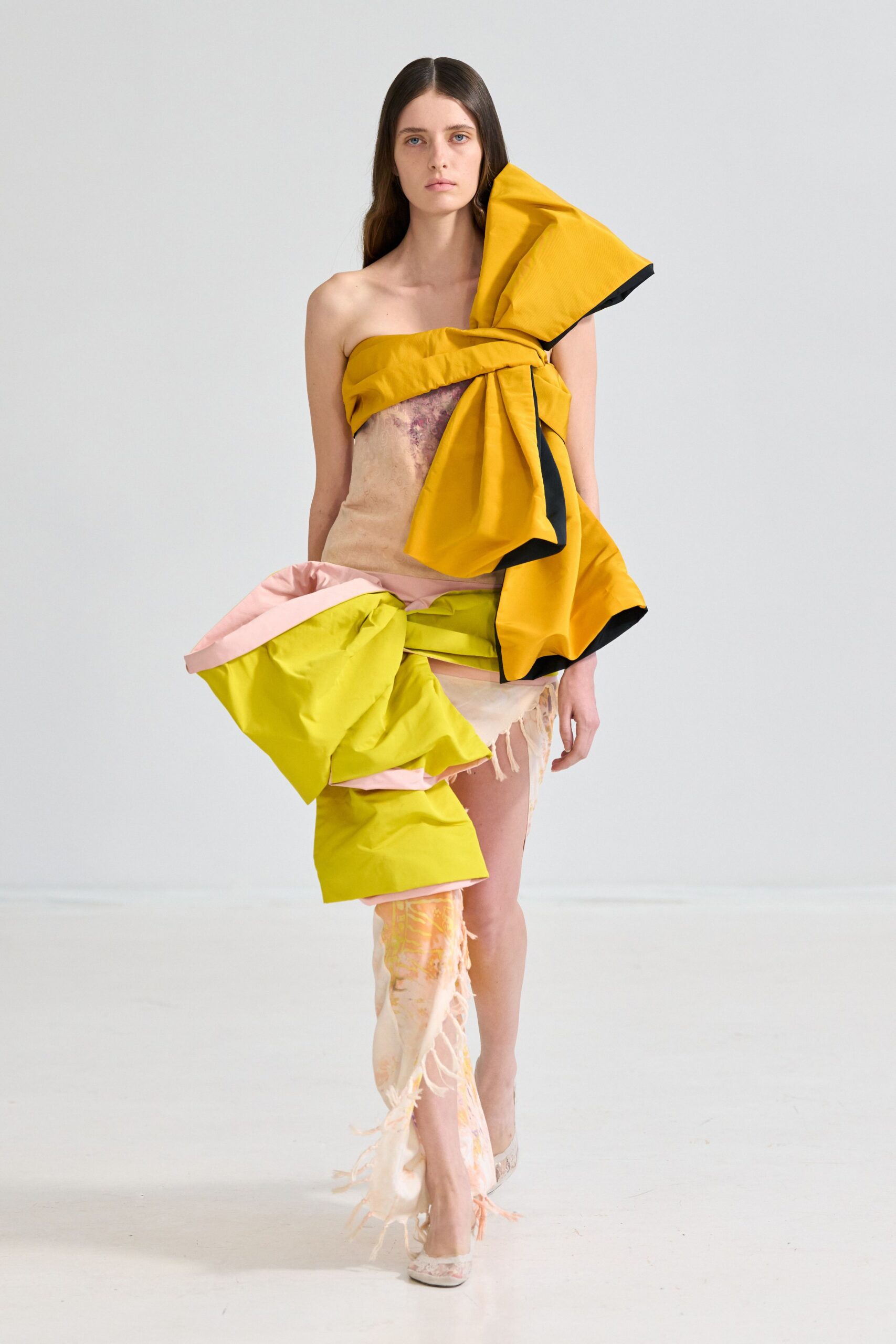 Asymmetric strapless dress wrapped in inflated marigold and citron bands, black lining flashing. Blush gauze panel hangs shredded at one thigh, watercolor stains ghosting through. Pale pumps in matching printed fabric. After a workshop where packing foam, muslin and paint never got put away.