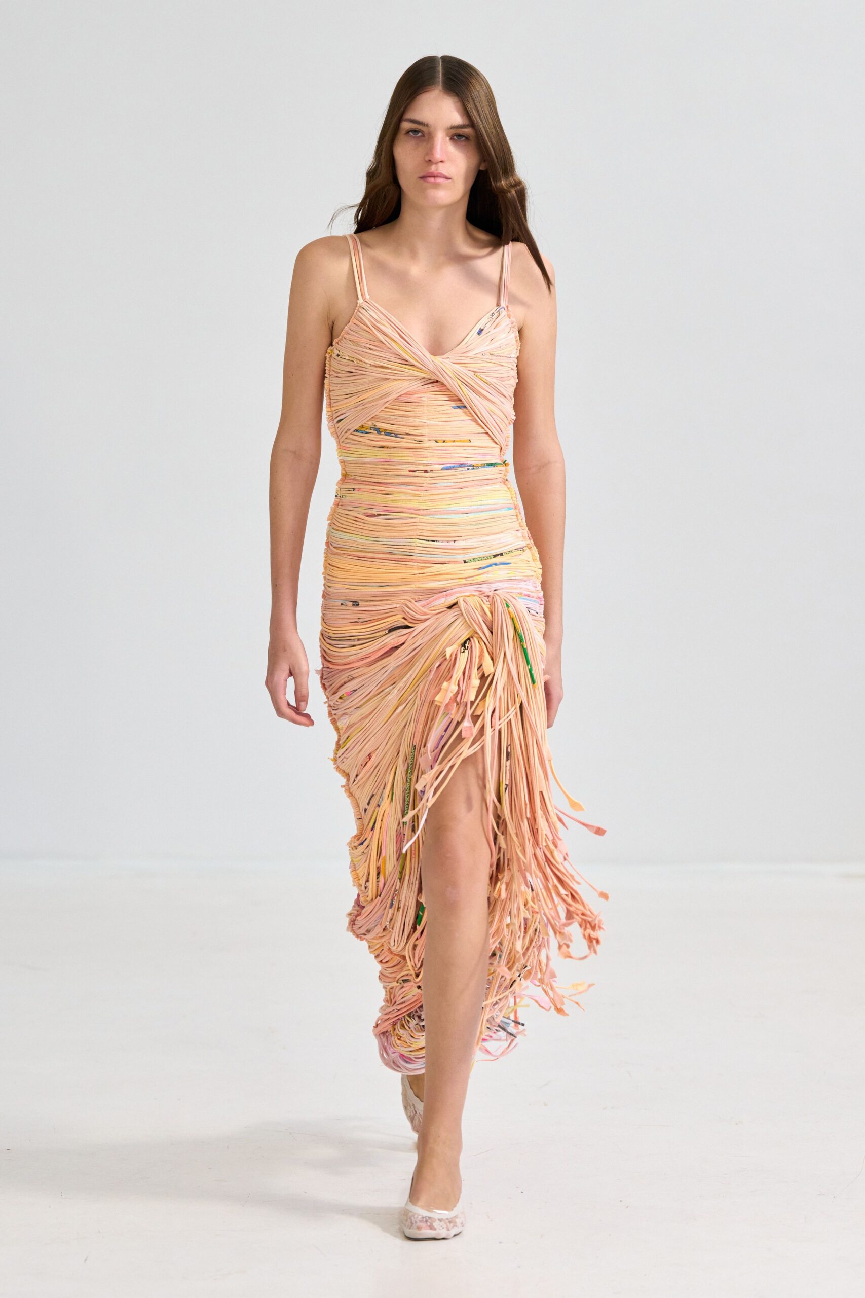Blush spaghetti-strap dress built from tightly wrapped jersey cords, exploding into dangling ends at the hip slit. Pale pointed flats with a cloudy marbled finish. Whatever survives the wedding after the cake table collapses.