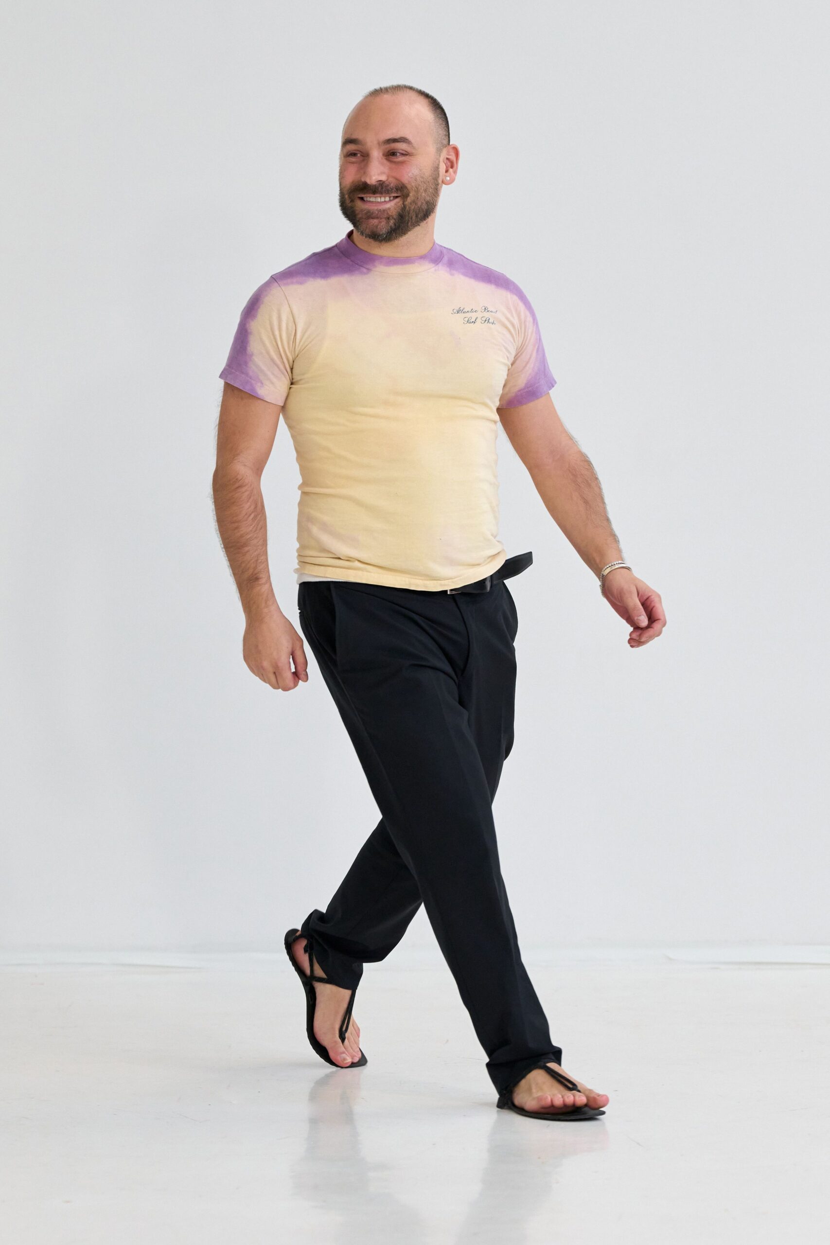 Fitted cream cotton tee, airbrushed lavender at the shoulders and sleeves. Black straight trousers with a slim belt tongue jutting forward. Flat black thong sandals, barely more than straps. Thin silver bangle stack. Walking back from the beach bar before the pavement loses its heat.