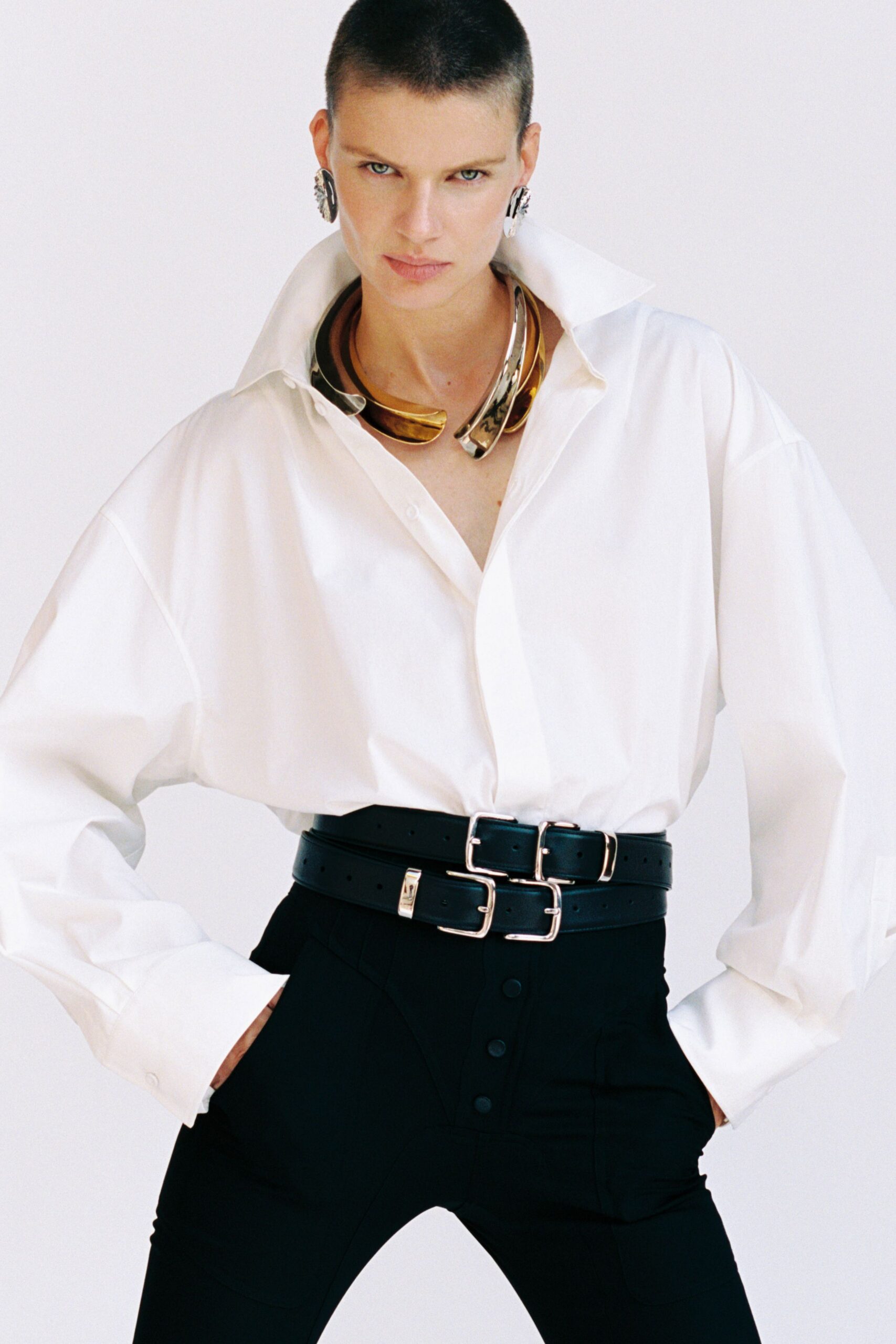 Silver shell earrings hugging both lobes. Wide mixed-metal collar, one end bent into a hooked tip. Oversized white cotton shirt with a hard popped collar. Double black leather belt stacked with bright square buckles. High-waisted black trousers with a buttoned placket. For taking over the meeting after the projector failed.