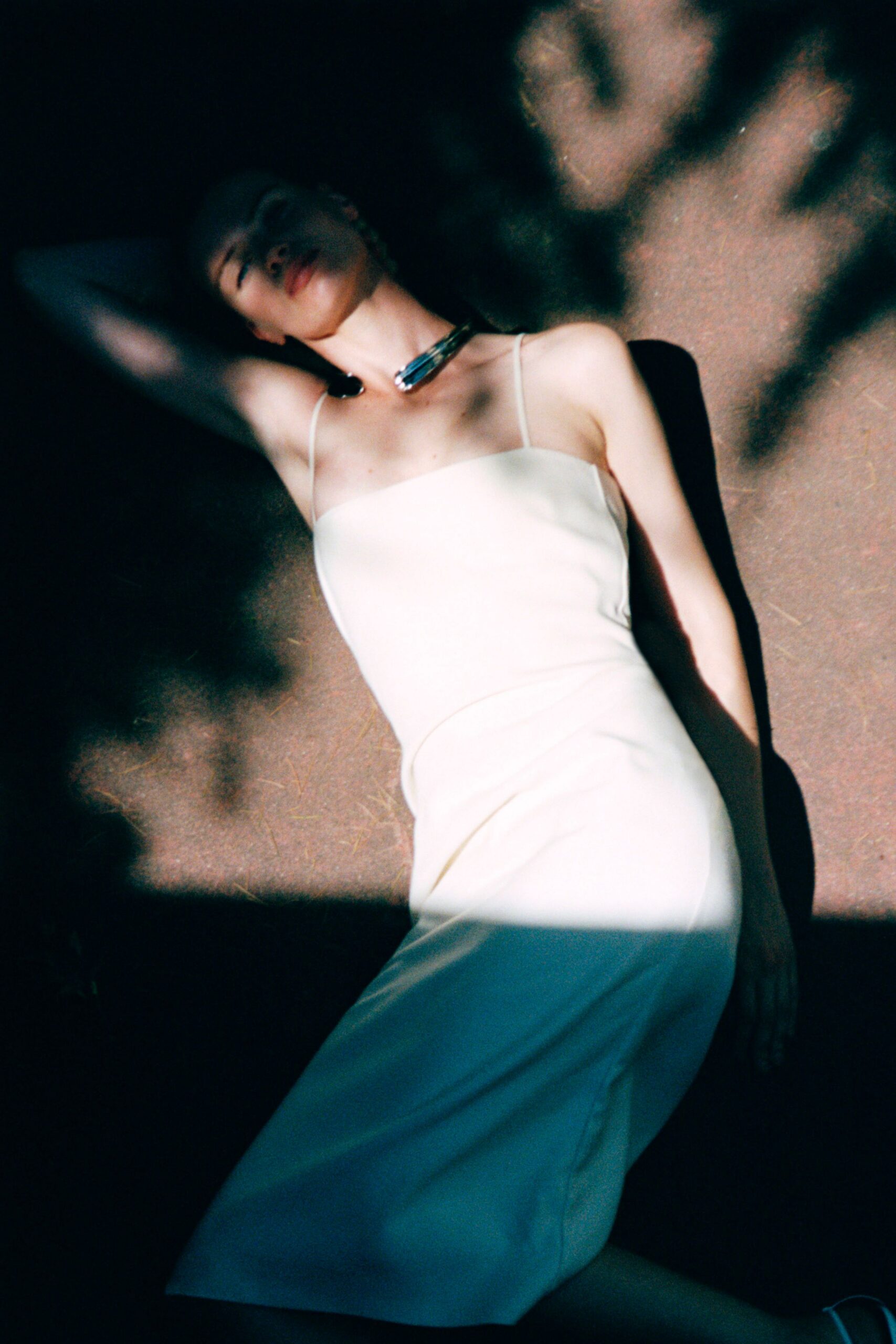 Ivory slip dress in dense crepe, straight neckline on filament straps. Rigid silver collar curving forward in two blunt glossy ends. Sun through a train window, halfway to a courthouse wedding.