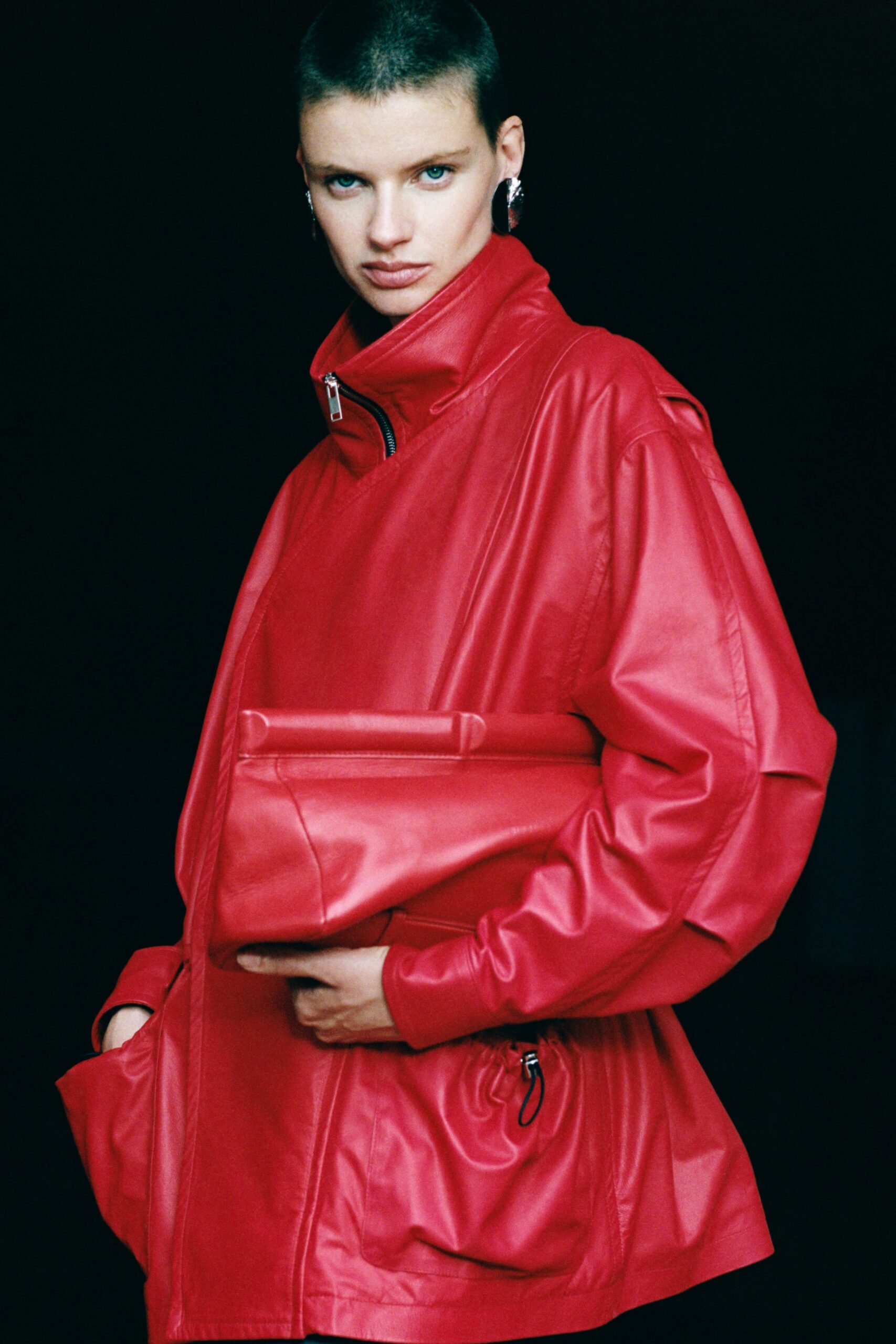 Cherry red leather jacket with a high zipped collar and dropped shoulders. Matching folded leather clutch tucked flat under the arm. Chunky silver earrings catching at the lobes. Red leather waistband cinched with a black cord toggle. Waiting at the ferry terminal after missing the earlier boat.