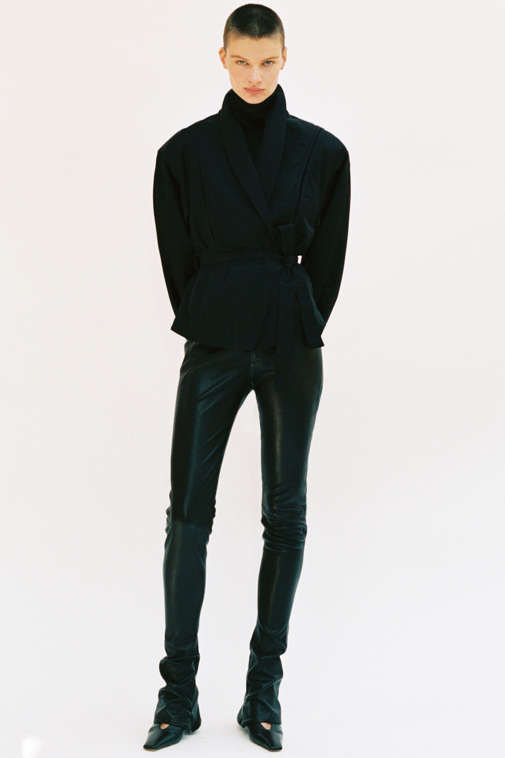 Black knit turtleneck rising close under the jaw. Inky wrap jacket with a shawl collar and tied side belt. Black leather leggings, glossy and creased at the knees. Pointed black boots with ruched shafts pooling at the ankle. For crossing an empty hotel lobby before sunrise checkout.