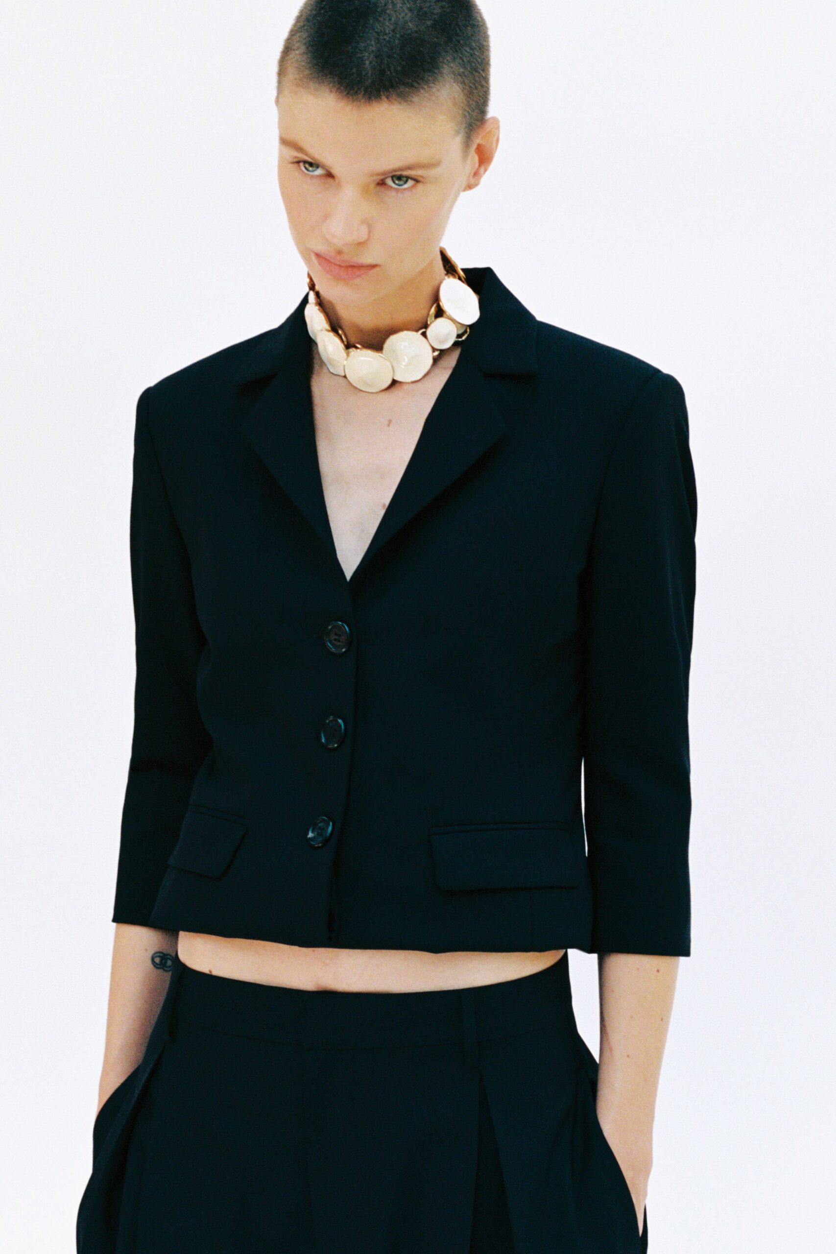 Chunky ivory disc necklace linked with warm gold hardware. Black cropped blazer with three glossy buttons and sharp notched lapels. High-waisted black trousers falling from a flat front into wide pleats. Museum cafe at noon, still wearing the necklace from last night.