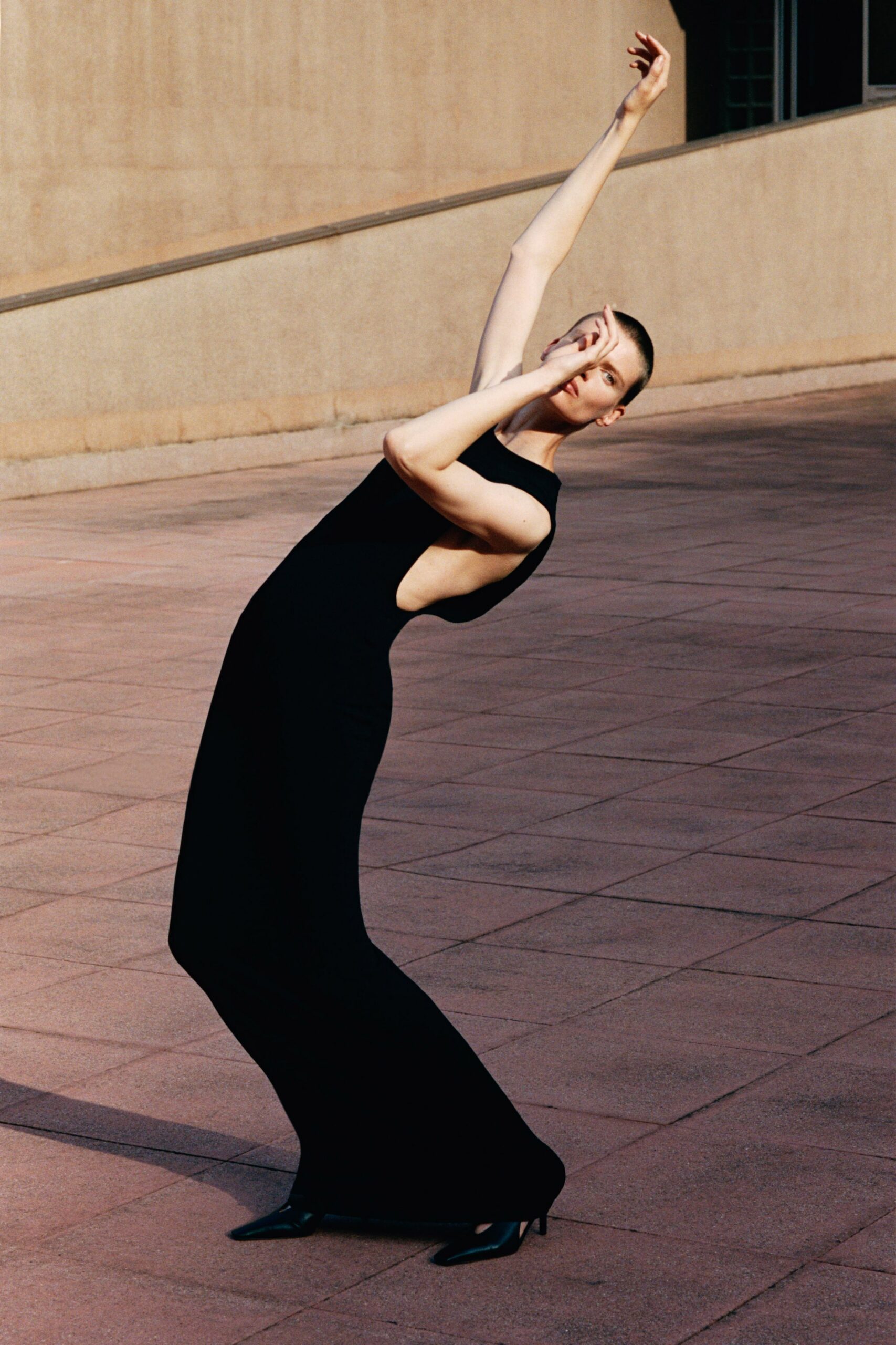 Black column dress in dense jersey, side cutout scooped nearly to the back. Pointed black pumps with low needle heels. Sunstruck civic plaza, nobody else around to see the pose.