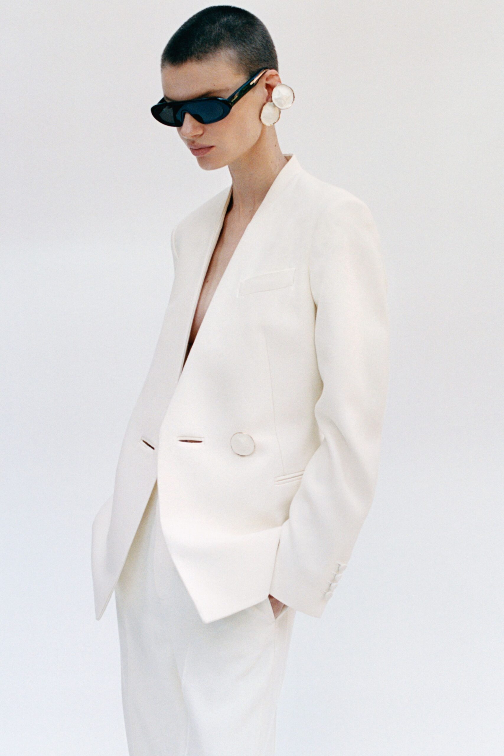 Narrow black wraparound sunglasses with thick glossy temples. Stacked white disc earrings, brushed like carved shell. Ivory collarless blazer cut deep, one covered button low on the waist. Wide ivory trousers falling in a flat uninterrupted line. In a white hotel corridor before the wedding guests start knocking.