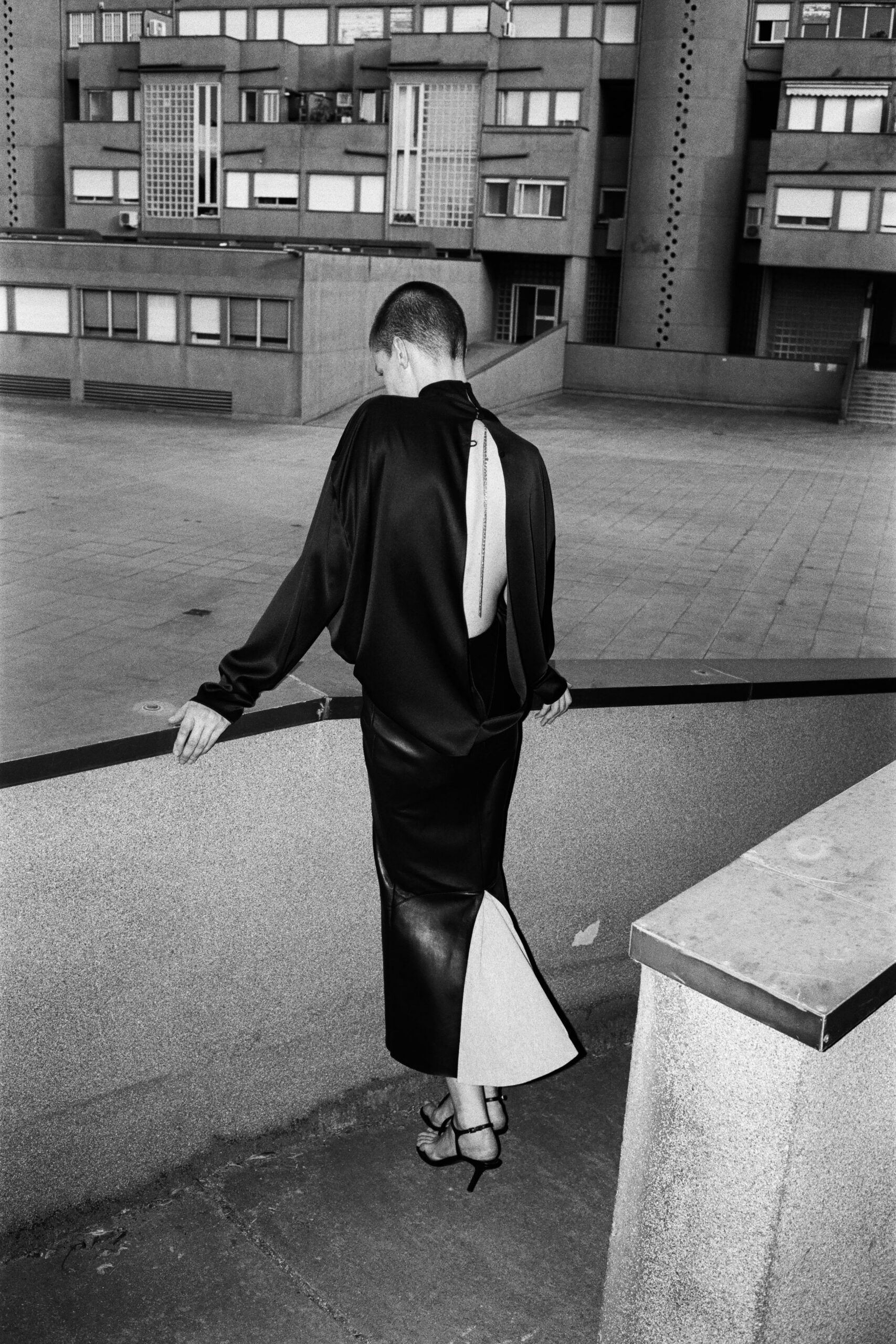 Black satin blouse cut open to the waist, chain running the spine. Black leather column skirt with a pale godet flaring from the back slit. Bare strappy heels with thin ankle buckles. Crossing a housing estate courtyard after midnight rain.