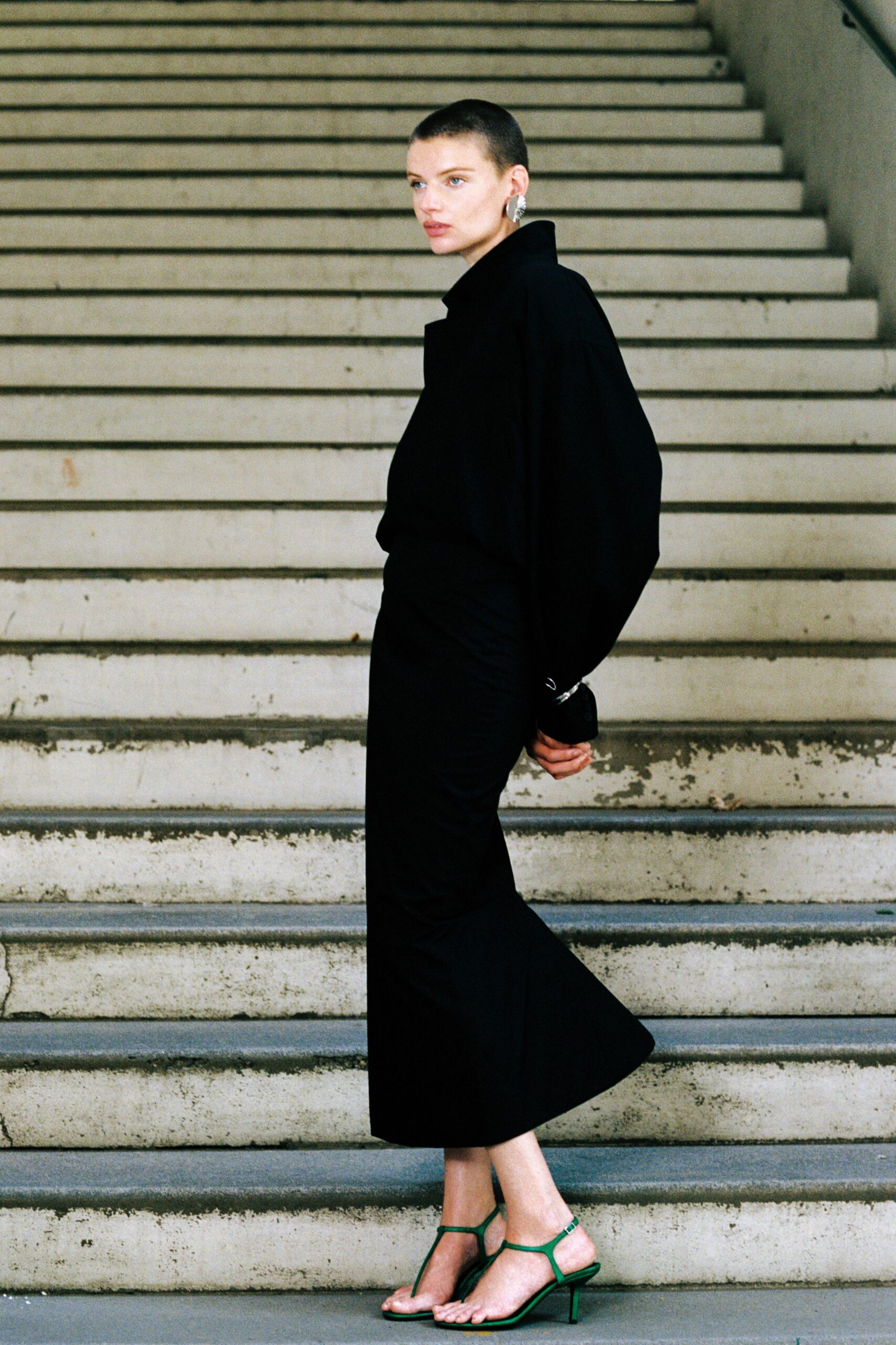 Black blouse with a high stand collar and billowed sleeves. Black column skirt, ruched close through the hip then loosening. Crinkled silver shell discs. Green thong sandals with spindly heels and squared soles. Crossing a civic stairwell after rain, still holding the last word.