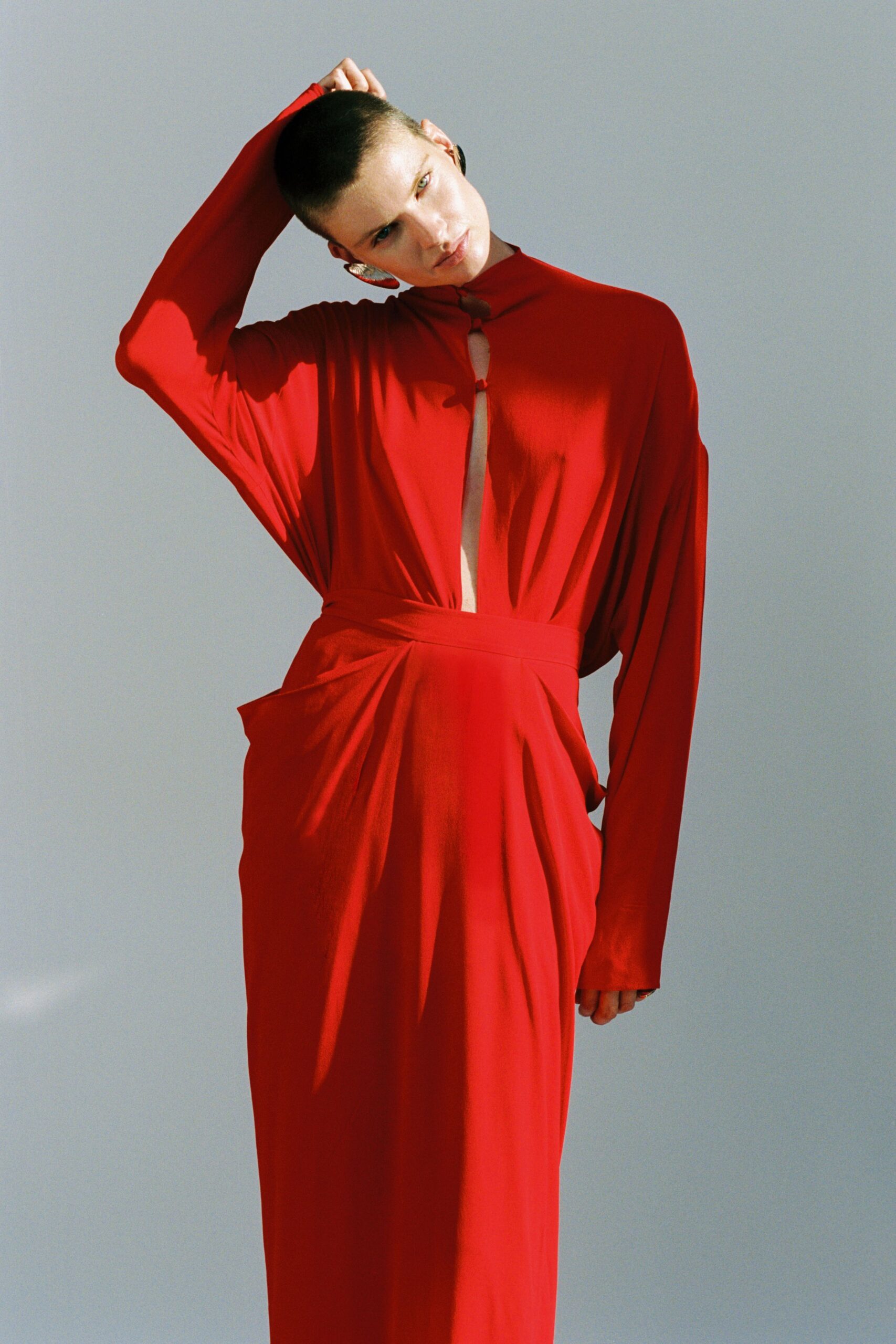 Tomato-red jersey column dress with batwing sleeves and looped slit fastening. Hammered silver discs catching beside the buzz cut. Waiting for the courthouse doors to open after lunch.