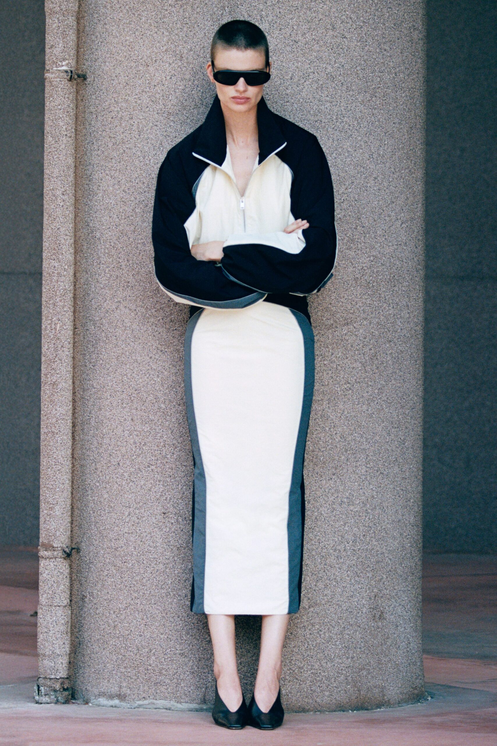 Black wraparound shield sunglasses. Cream jersey zip top with black raglan sleeves and a standing collar. Column skirt in matching cream jersey, slate side panels lengthening the line. Black pumps with a squared throat. Waiting for the municipal building doors to unlock after lunch.