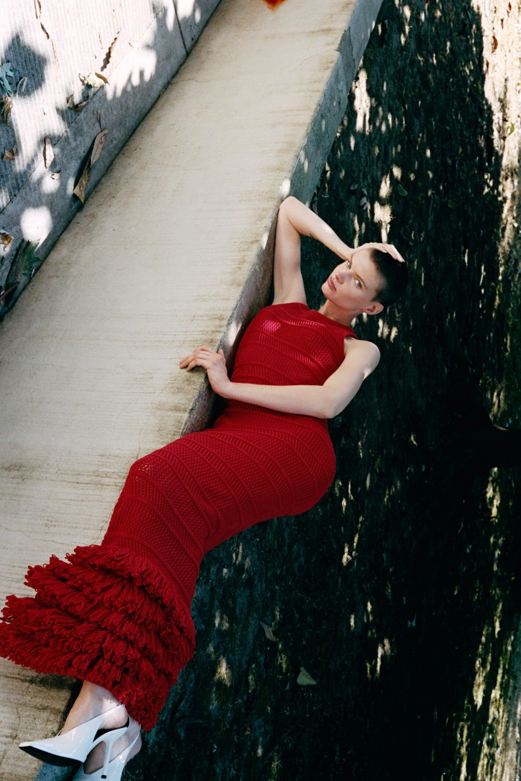 Scarlet open-knit column dress, banded through the body with a shaggy fringe hem. White patent slingbacks with knife-point toes. Caught in the dry concrete ditch behind a hotel after noon.