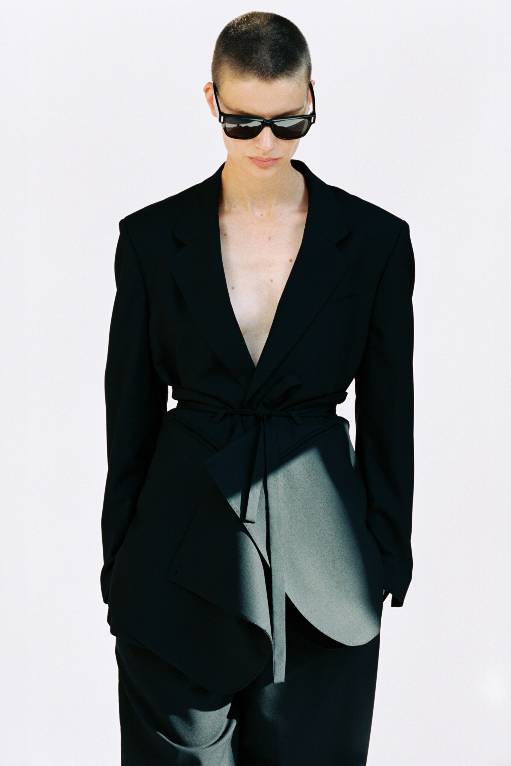 Glossy black rectangular frames with thick temples. Black tailoring worn bare, wrapped tight with a self-tie waist. Black straight trousers in a fluid suiting drape. Midmorning hotel checkout, sunlight slicing across the lobby floor.