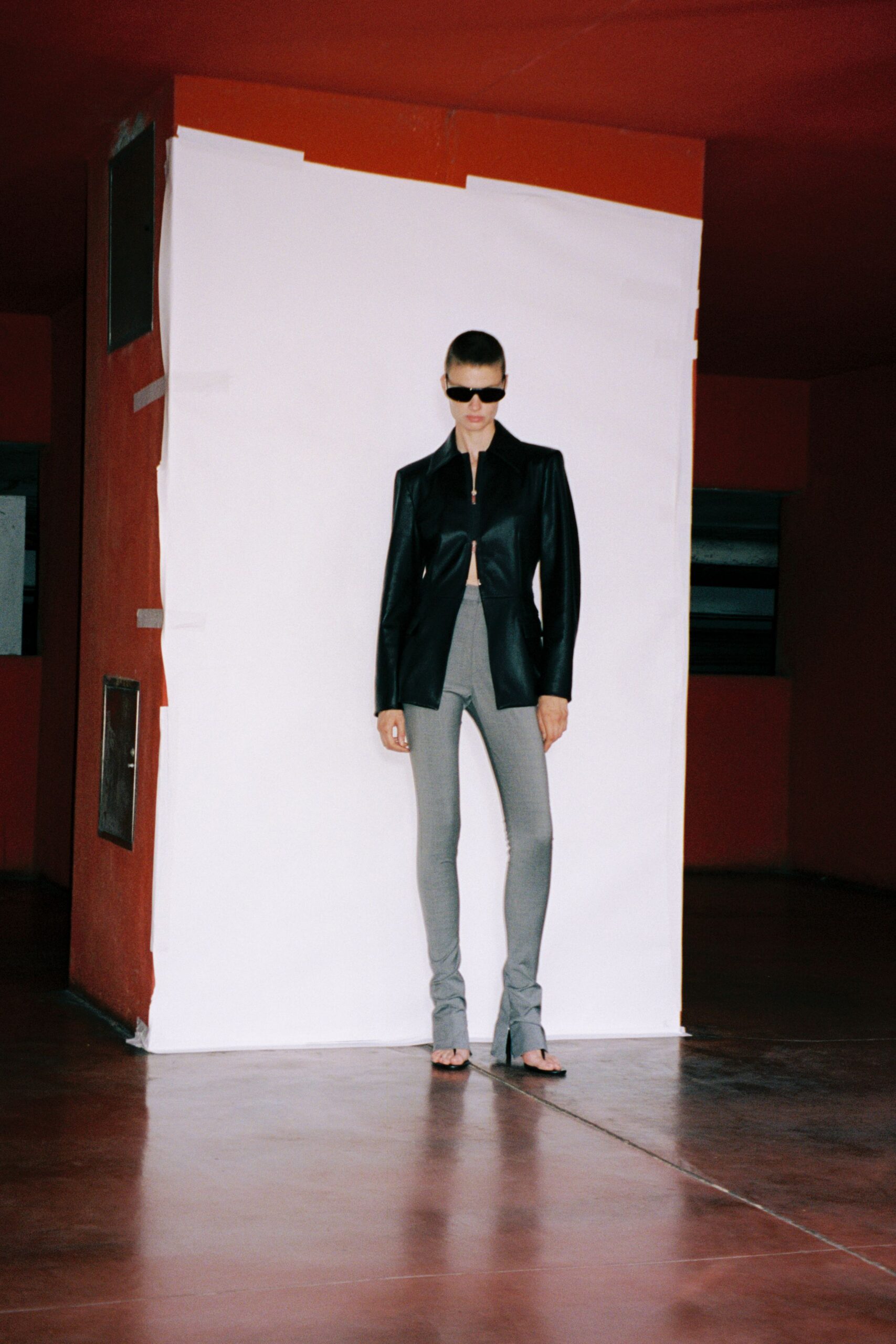 Black wraparound sunglasses, narrow and opaque. Black leather jacket, cutaway front held by a tiny top zip. Pale grey stirrup leggings, split over the instep. Barely there black thong sandals. Crossing an empty office atrium before the air conditioning starts.