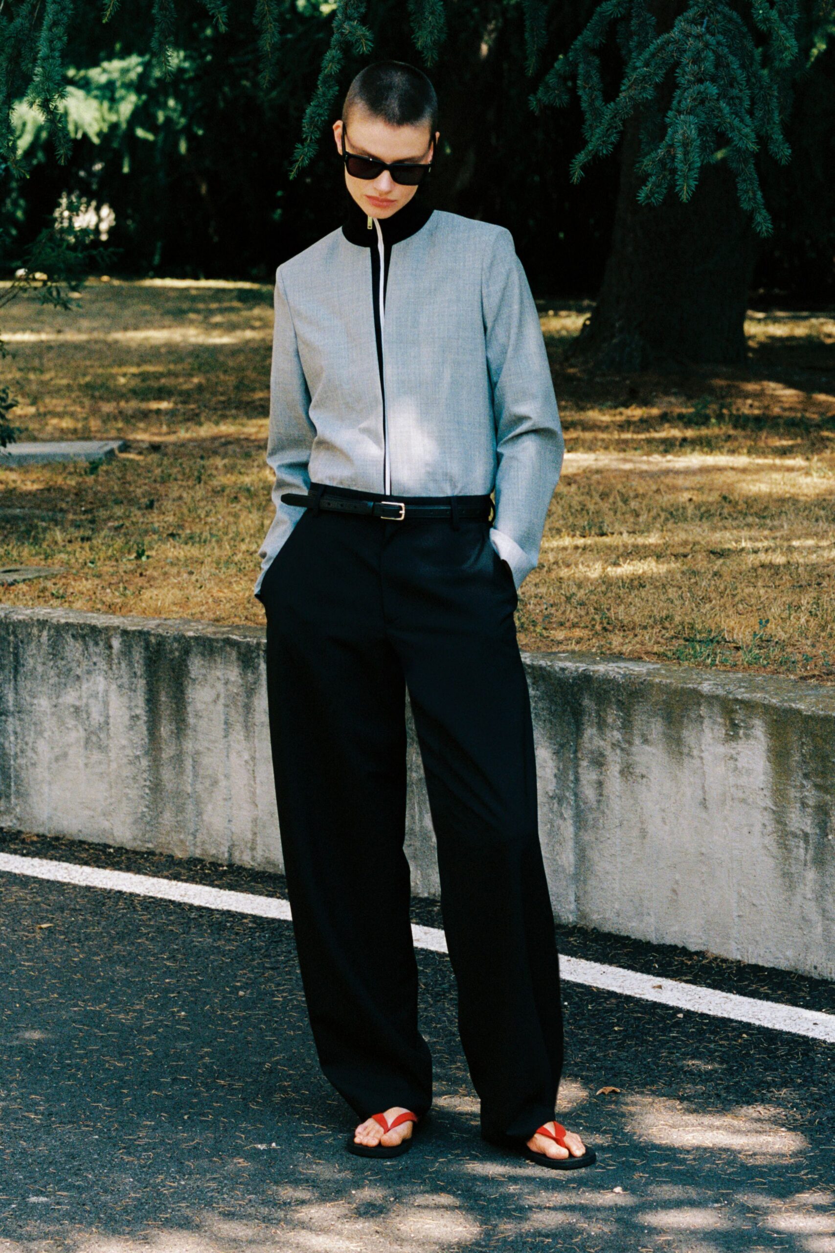 Wide black rectangular frames. Pale grey zip-front jacket with a tall black stand collar. Slim black leather belt with a small silver buckle. Black wide-leg trousers breaking softly over the foot. Black rubber soles with bright red thong straps. Crossing the botanical garden car park before the heat fully settles in.