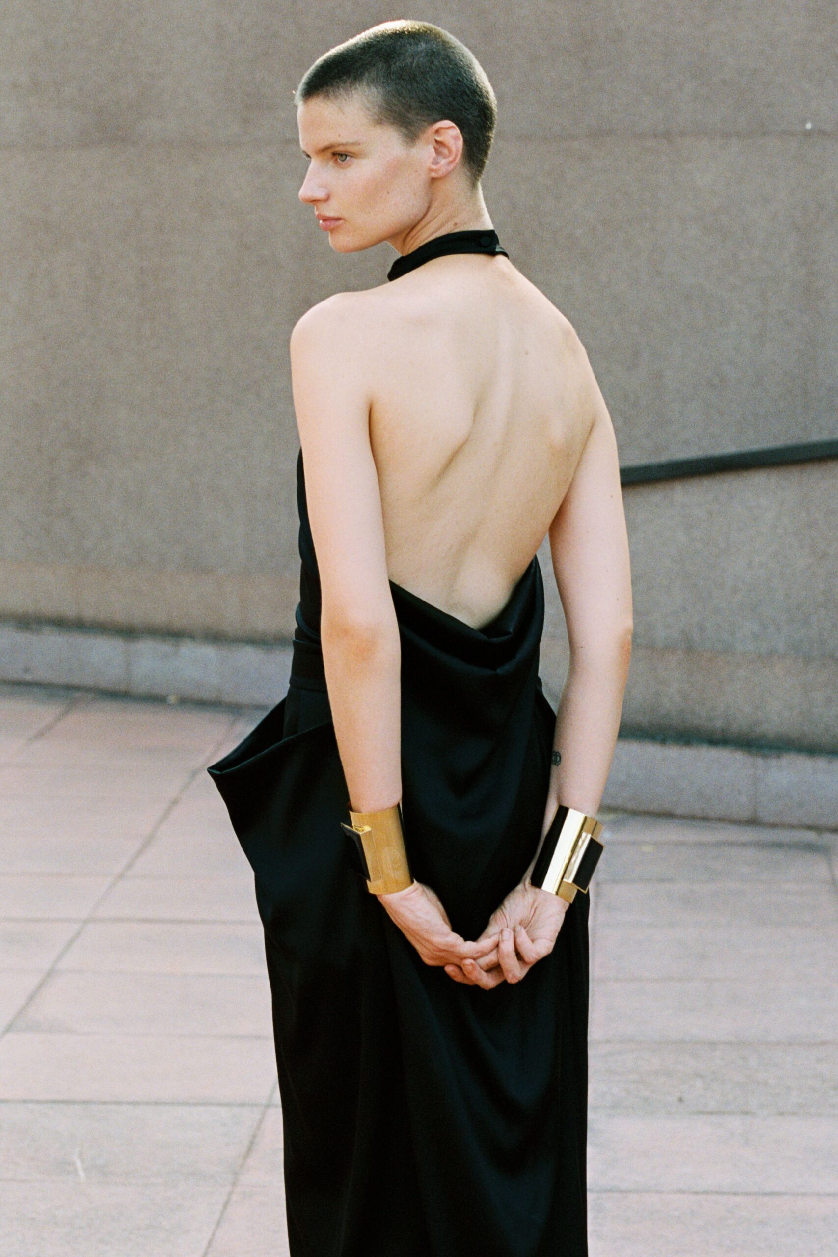 Black satin halter gown with a bare back and folded peplum hip. Wide mirror-bright gold cuffs, one angled open at the wrist. Museum steps after dusk, stone still warm underfoot.