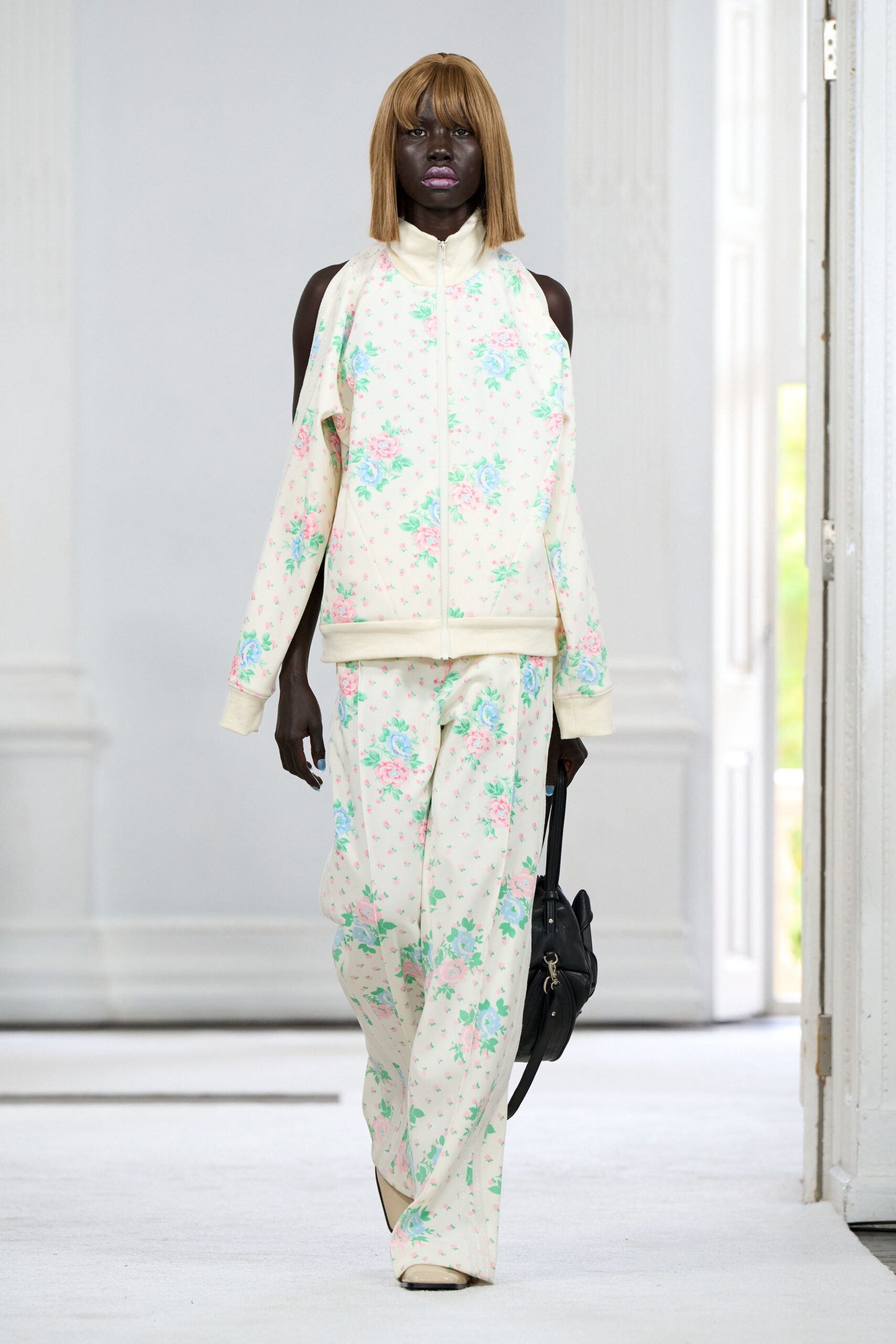Cream jersey track jacket in cabbage-rose print, shoulders sliced open beneath the funnel neck. Matching loose track pants, floral scattered over a soft drape. Black shoulder bag with dangling silver rings. Square-toe cream boots. Walking back from the corner shop with peonies wrapped in yesterday's paper.