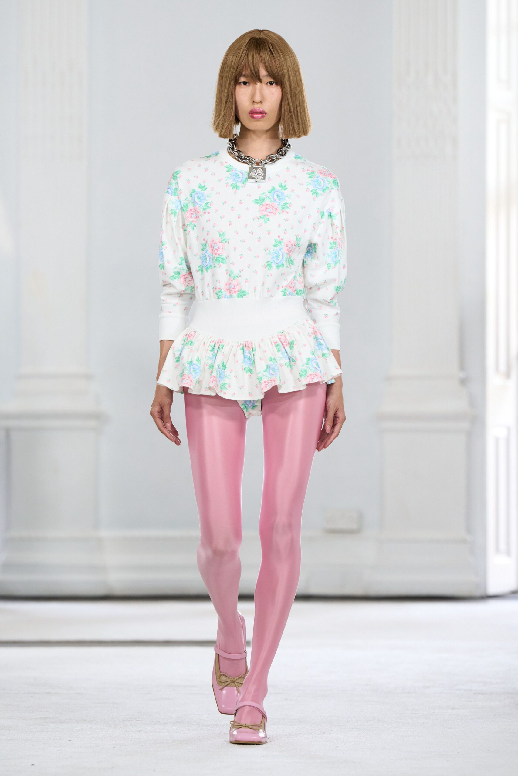 White jersey top printed with pastel cabbage roses, dropped into a peplum. Heavy silver chain with a square padlock clasp. Glossy bubblegum-pink tights pulled seamlessly into the shoe. Square-toe pink patent Mary Janes with a small beige bow. Walking across a hotel ballroom before the bridal fair opens.