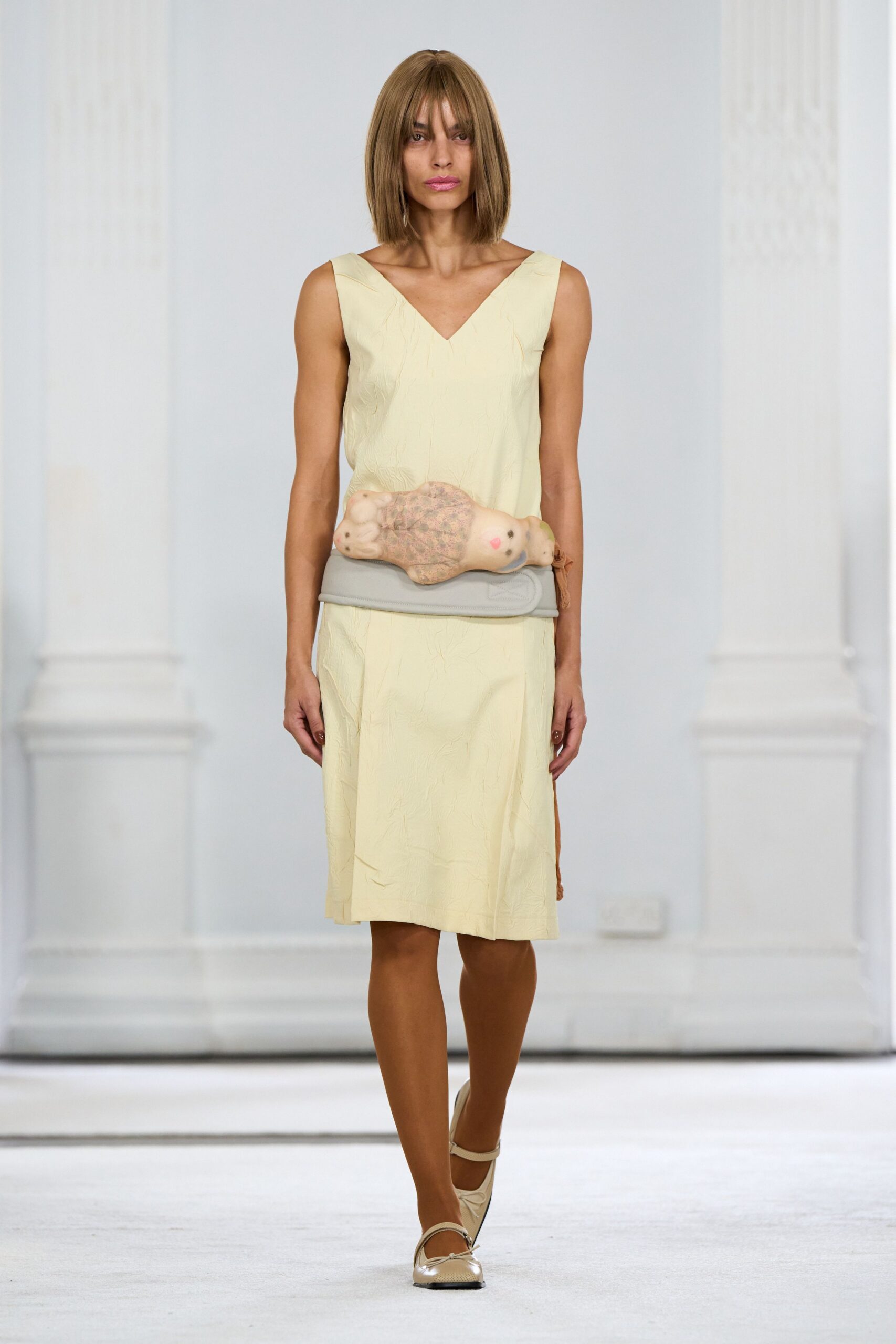 Pale butter sleeveless dress in lightly crinkled cloth, V-neck and calf slit. Pale grey padded belt with a stuffed doll strapped across the front. Cream Mary Jane flats with a tiny bow at the toe. For carrying something breakable through a silent house after midnight.
