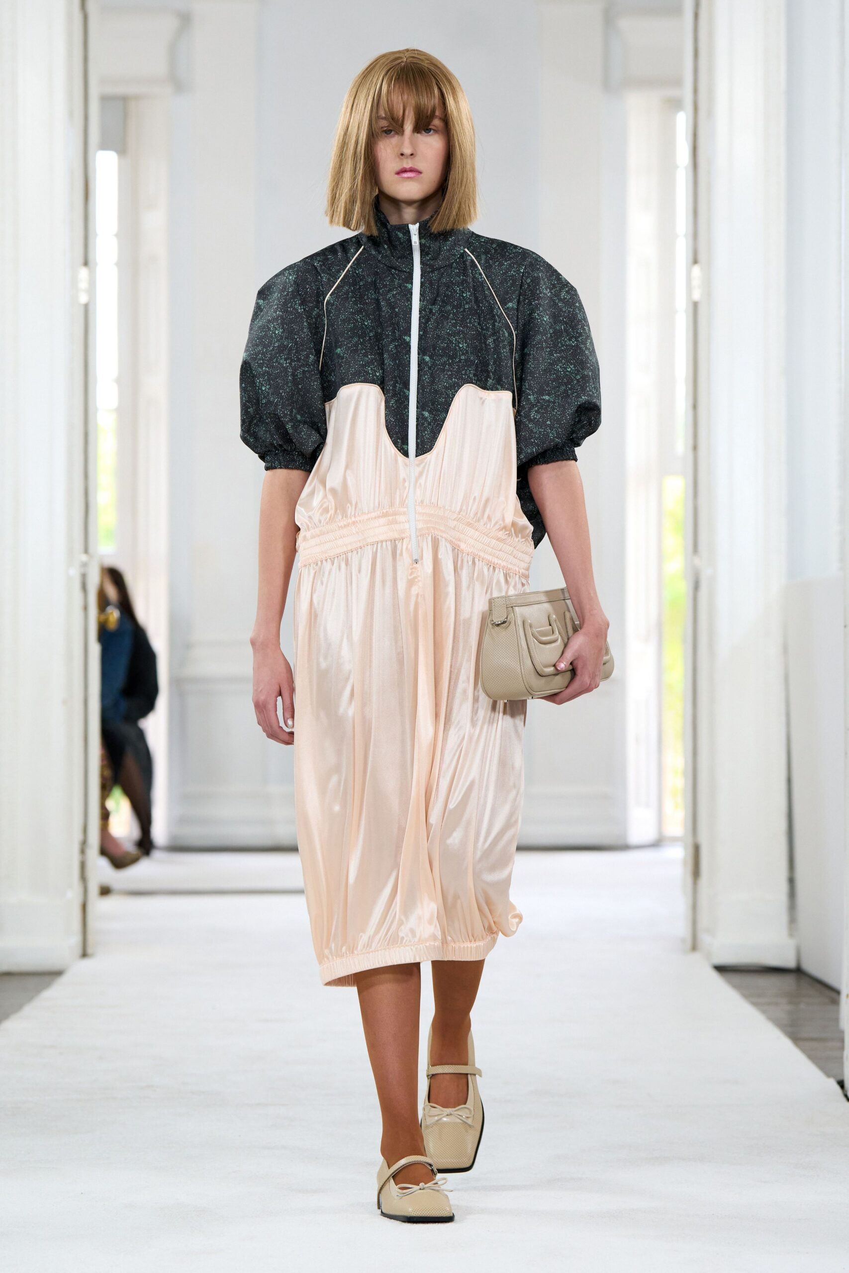 Black speckled zip jacket with puffed sleeves and pale piping. Blush satin dress gathered under a scalloped empire seam. Cream pebbled clutch with a tiny outer pocket. Square-toe cream flats, one bow slightly skewed. Walking the long hotel corridor before breakfast service starts.