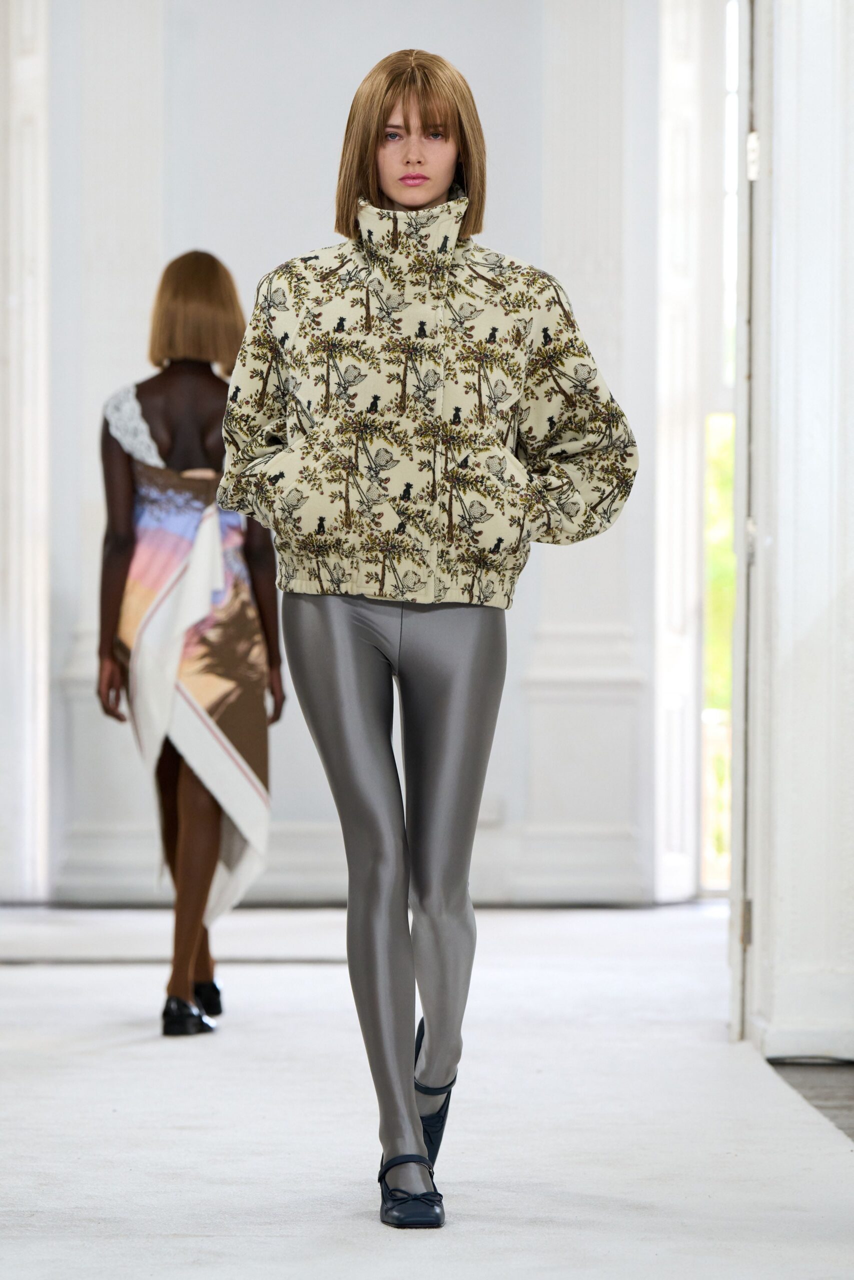 Cream quilted puffer printed with birds in climbing brown branches, funnel collar zipped high. Pewter stretch leggings with a wet-looking sheen. Black square-toe Mary Janes with a tiny bow. Walking the gravel path between aviaries before the grounds open.