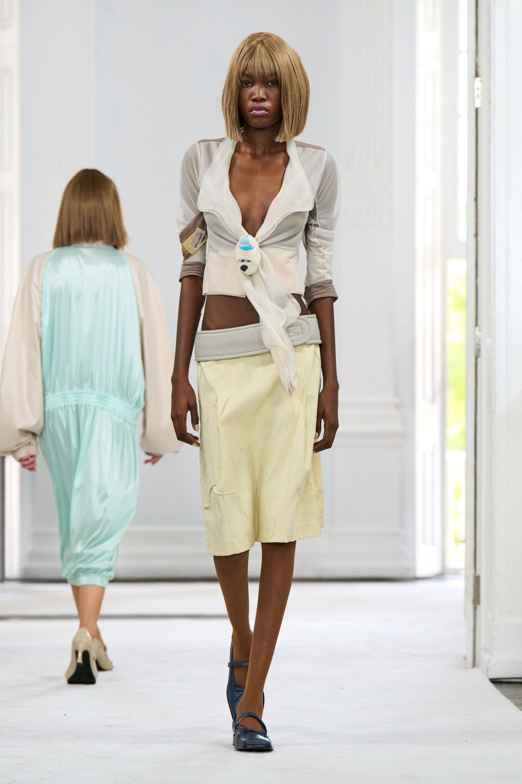 Sheer ivory blouse with a plunging scarf front pinned by a plush bear face. Pale butter knee-length skirt in crinkled satin, topped with a padded grey yoke. Navy Mary Jane flats. Crossing a sun-bleached corridor with a toy charm still hanging on.