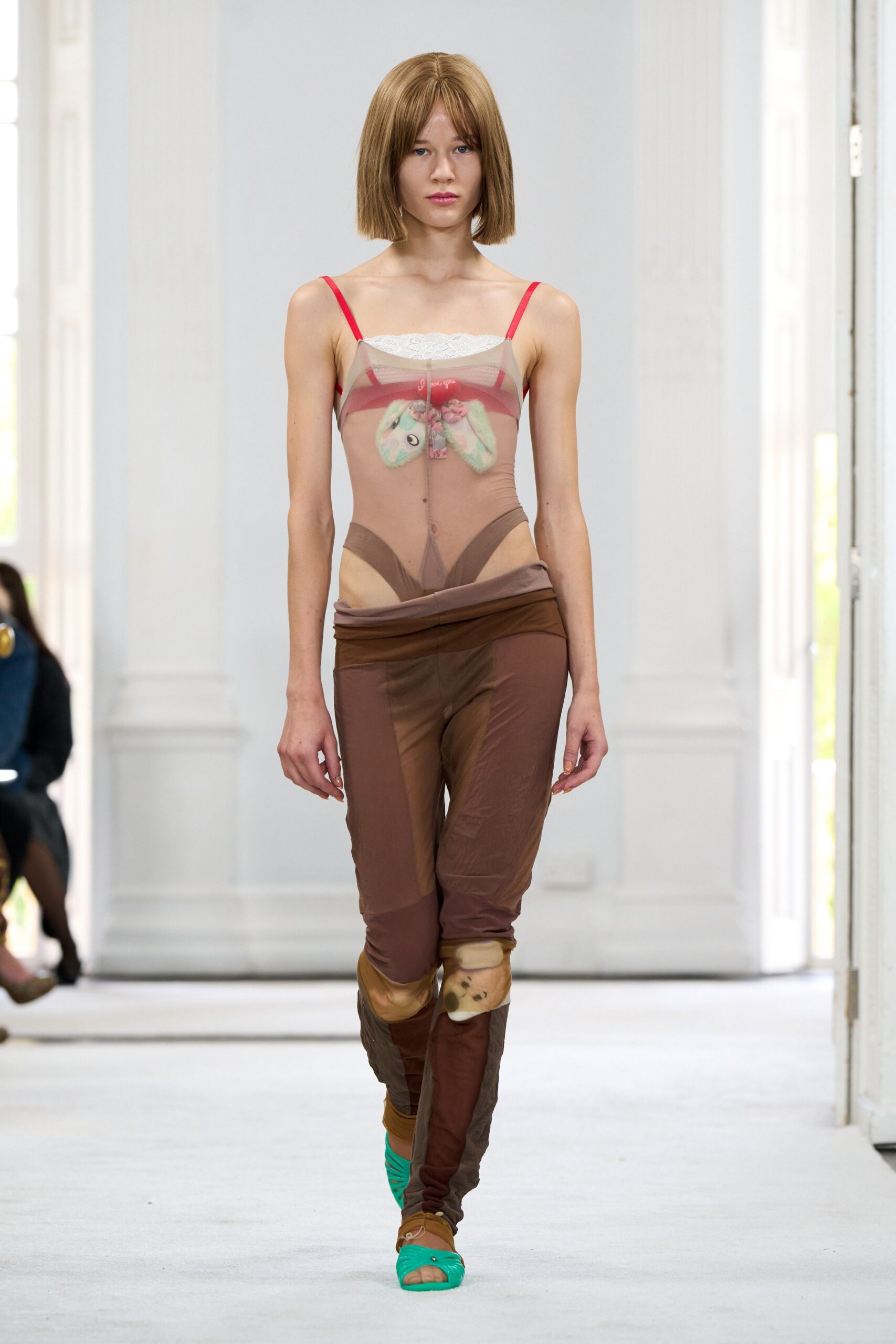 Taupe sheer mesh slip top, white lace peeking above red straps. Red bralette with a pastel stuffed bunny pinned at the chest. Brown stretch-mesh leggings, waistband rolled low and one knee patched plush. Bright aqua jelly sandals. For buying cough drops at a beach town pharmacy after sunset.