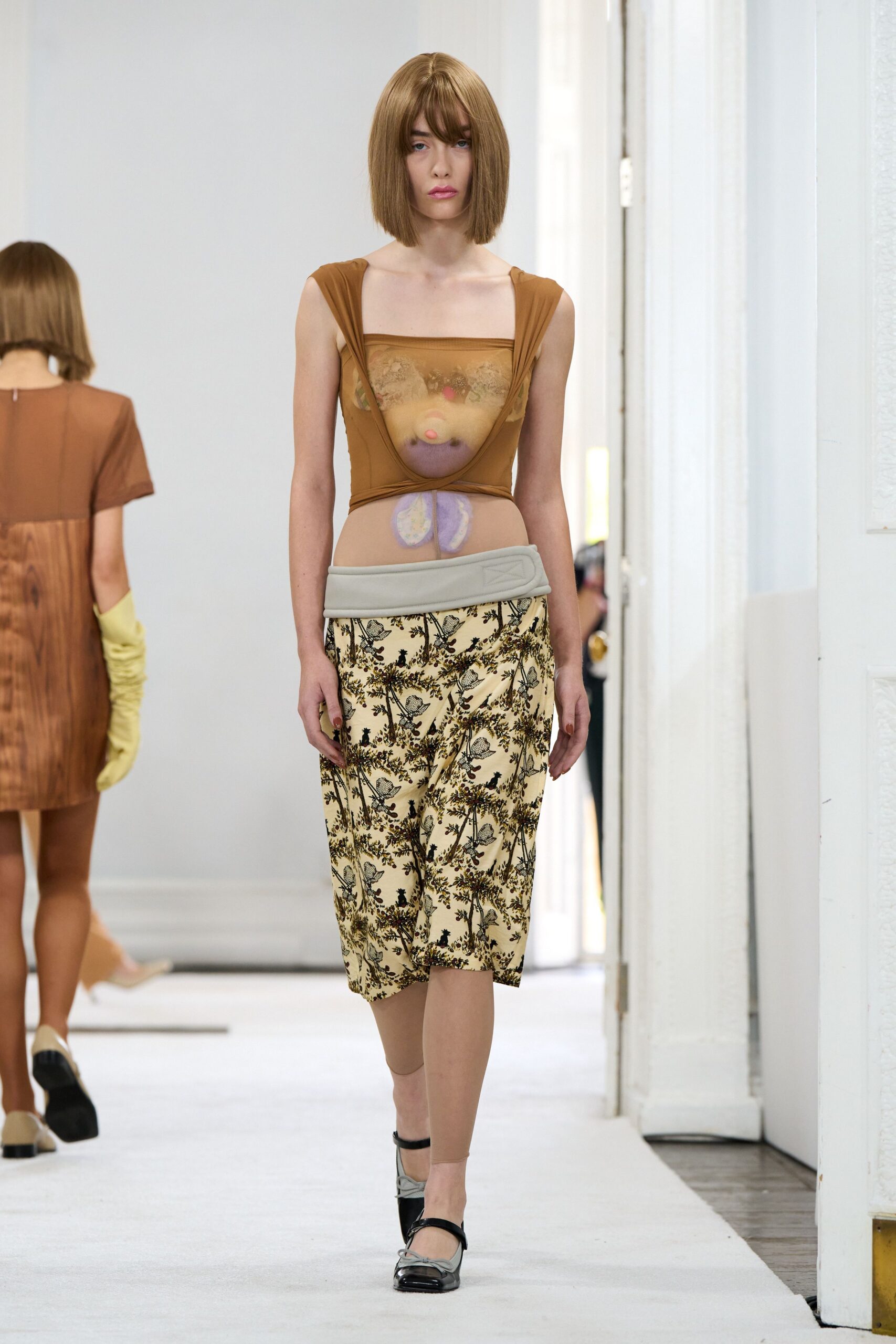 Tan mesh top draped into a deep U, with a stuffed bear trapped beneath. Cream knee-length skirt in a black woodland print, folded into a low padded waistband. Single nude stocking on one leg. Two-tone square-toe flats with thin ankle straps. For crossing a museum lobby carrying something strange under your shirt.